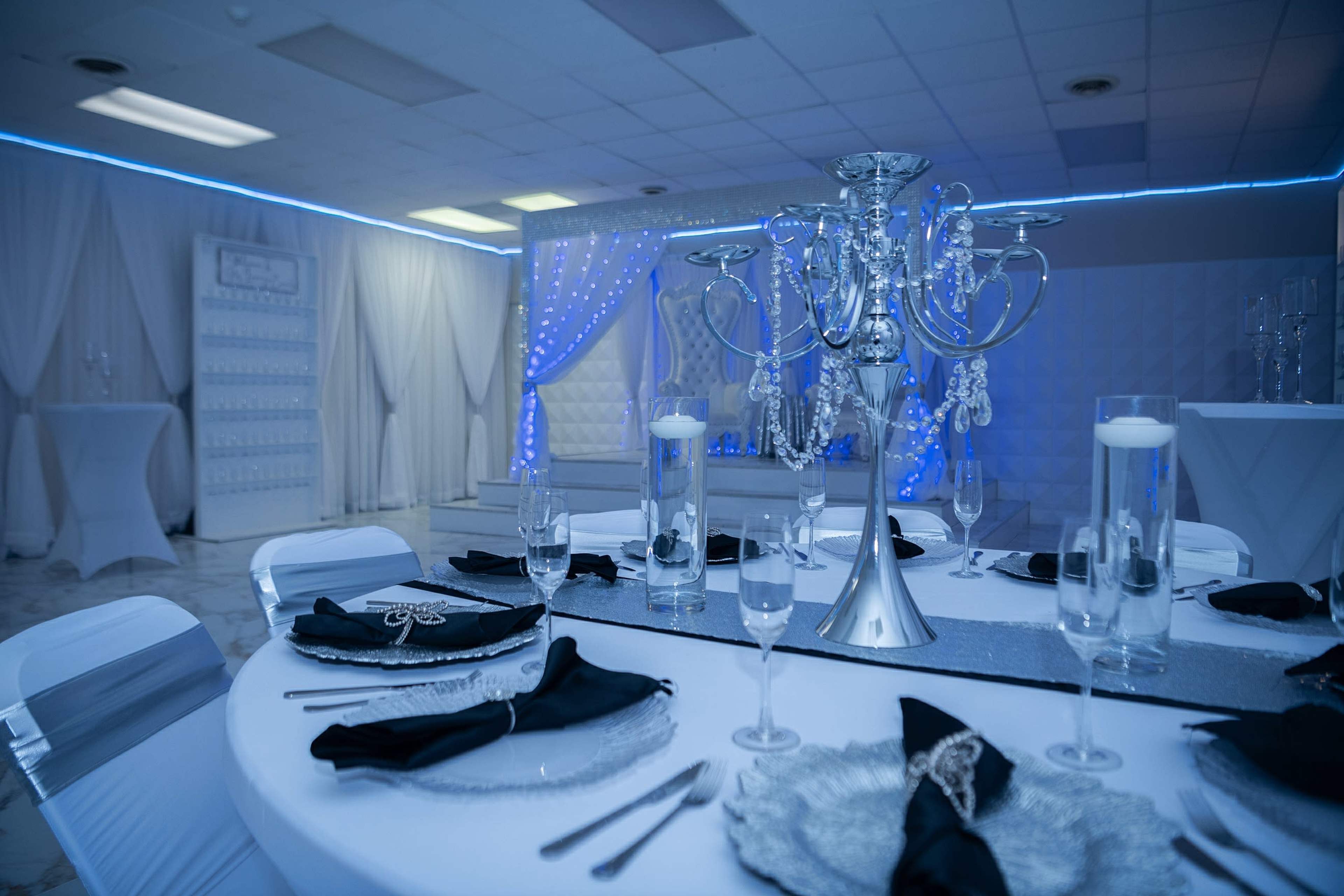 A elegantly decorated dining area with a round table set with black napkins, silver tableware, and a large silver candelabrum as the centerpiece, surrounded by white chairs and blue lighting.