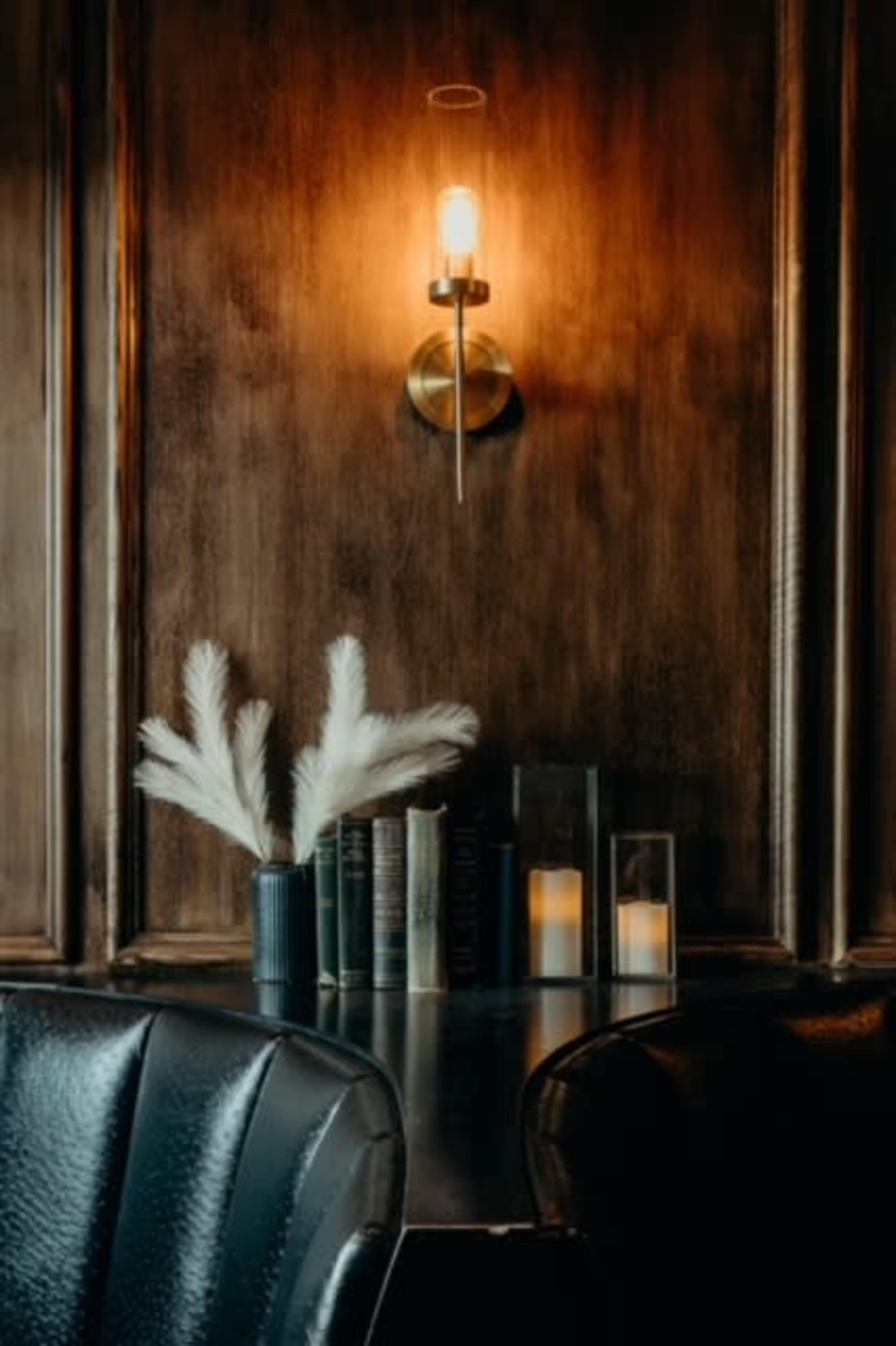 A cozy interior features a dark wooden wall, a wall-mounted light fixture, a collection of books, candles, and decorative dried plants on a table.