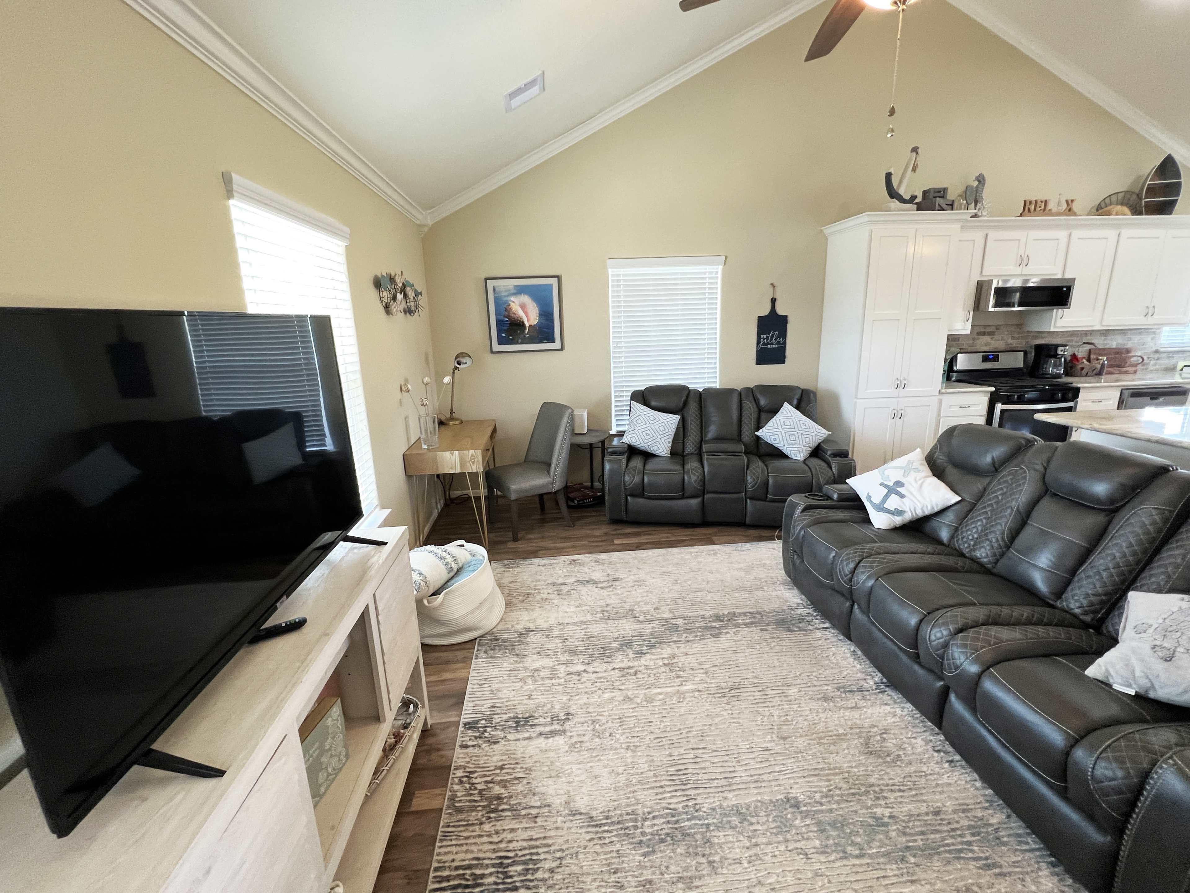 The living room features a large flat-screen television, two reclining sofas with decorative pillows, and a desk positioned near windows, set on a patterned area rug.