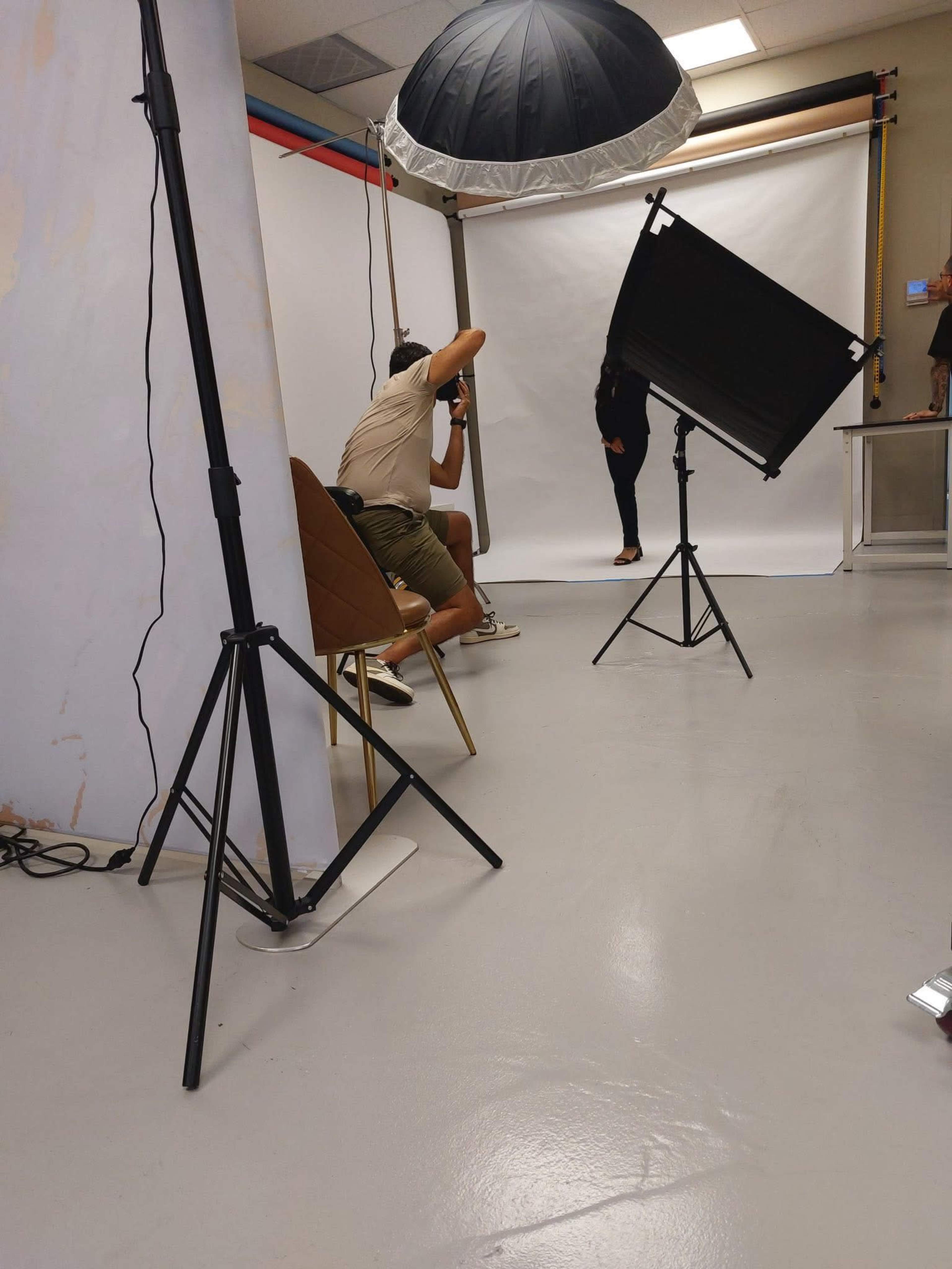 A photographer is taking pictures of a model in front of a plain white backdrop in a studio setting.