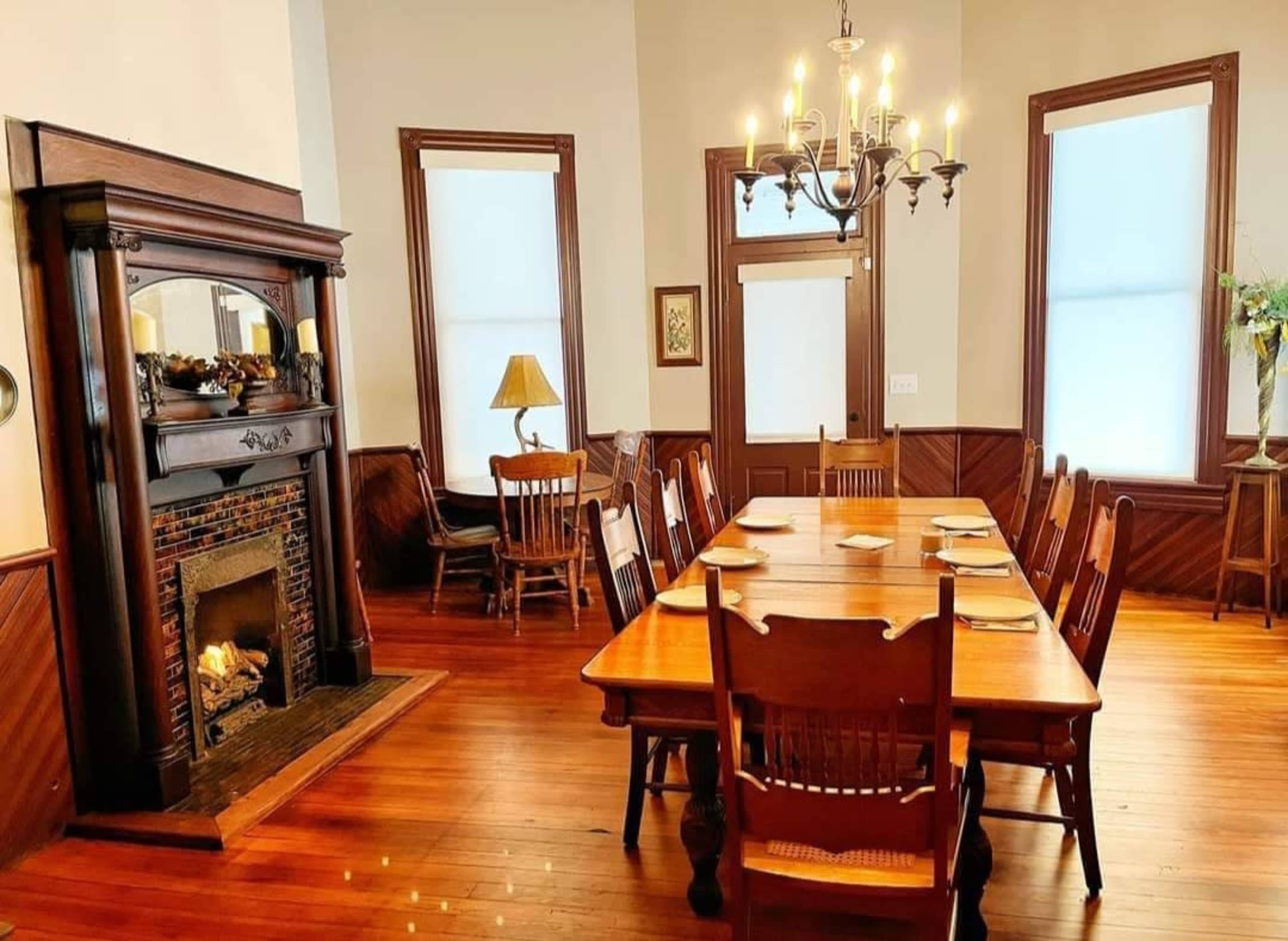 A spacious dining room features a long wooden table surrounded by chairs, with a fireplace and windows allowing natural light.