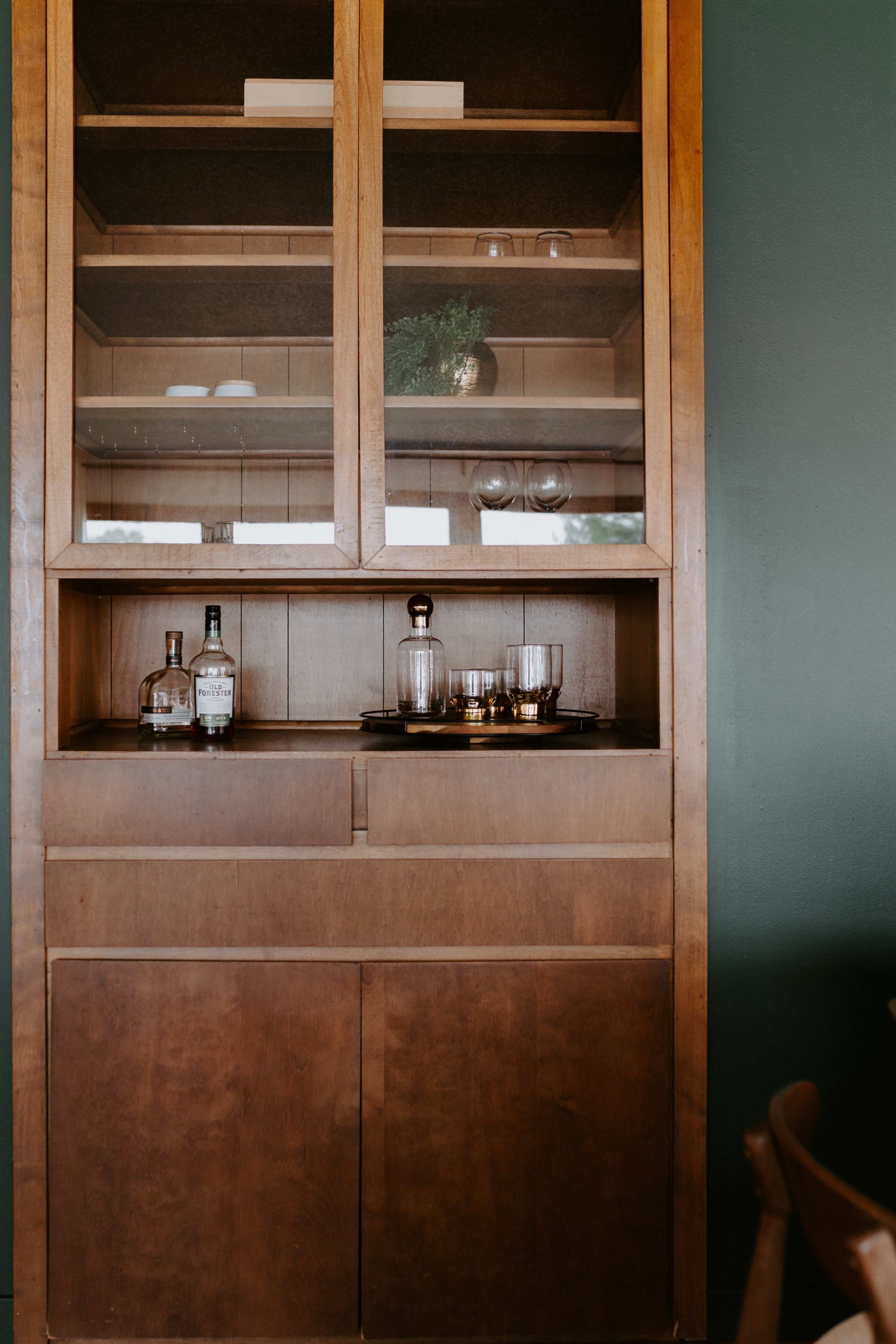A wooden cabinet with glass shelves displays drinkware and bottles, set against a green wall.