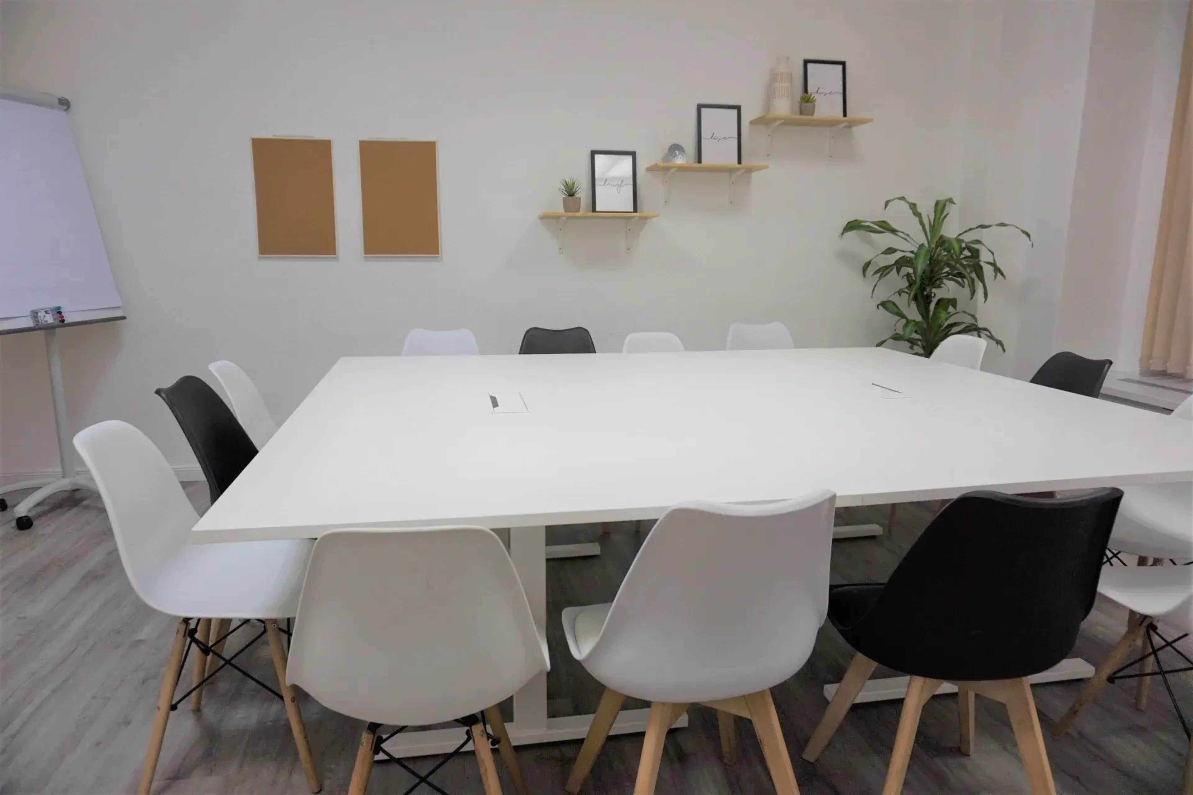 A large, white conference table is surrounded by a mix of black and white chairs in a minimalist meeting room with a plant and framed decorations on the walls.