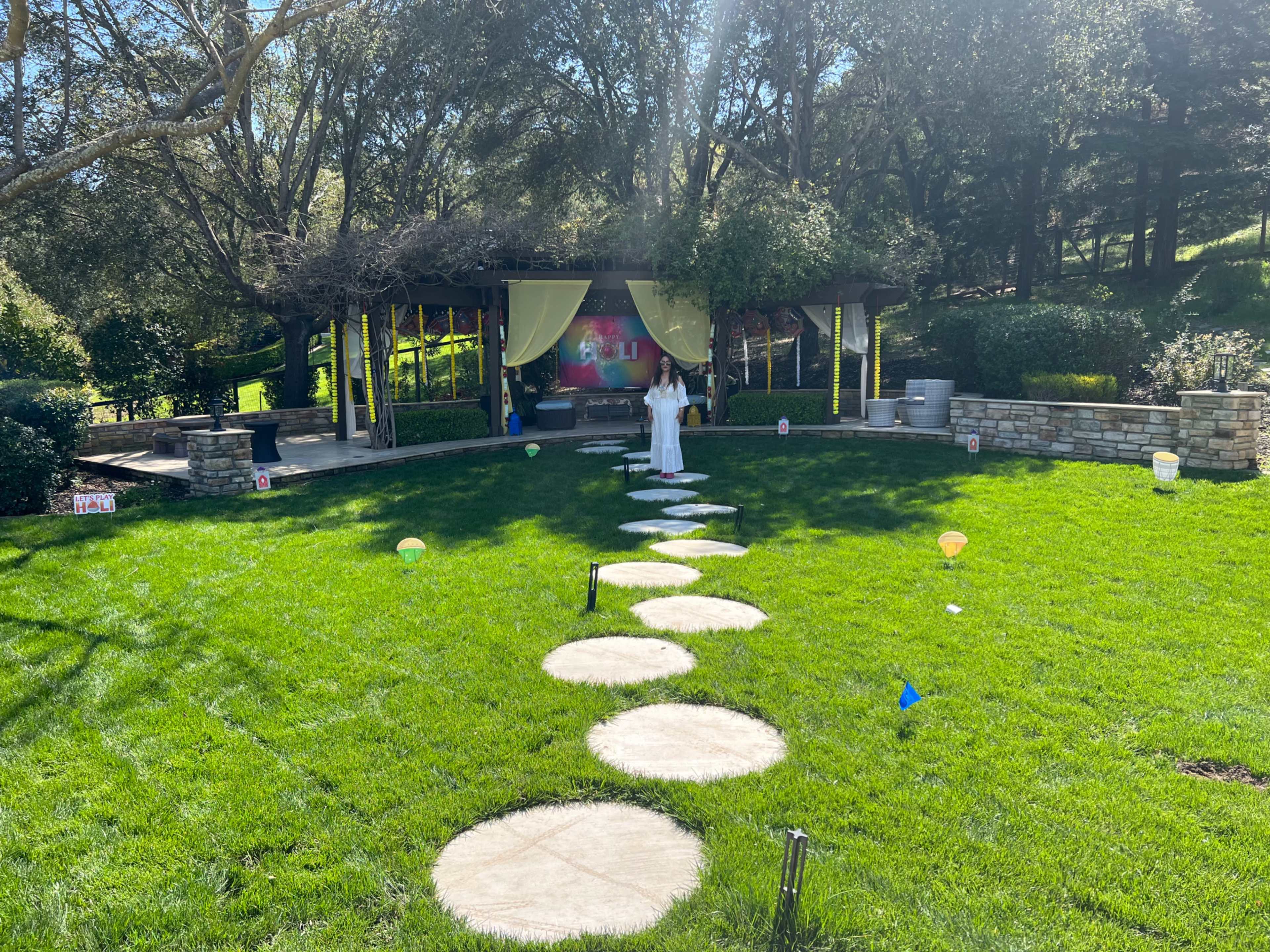 A woman in white stands on a stone pathway leading to a gazebo in a grassy area surrounded by trees.