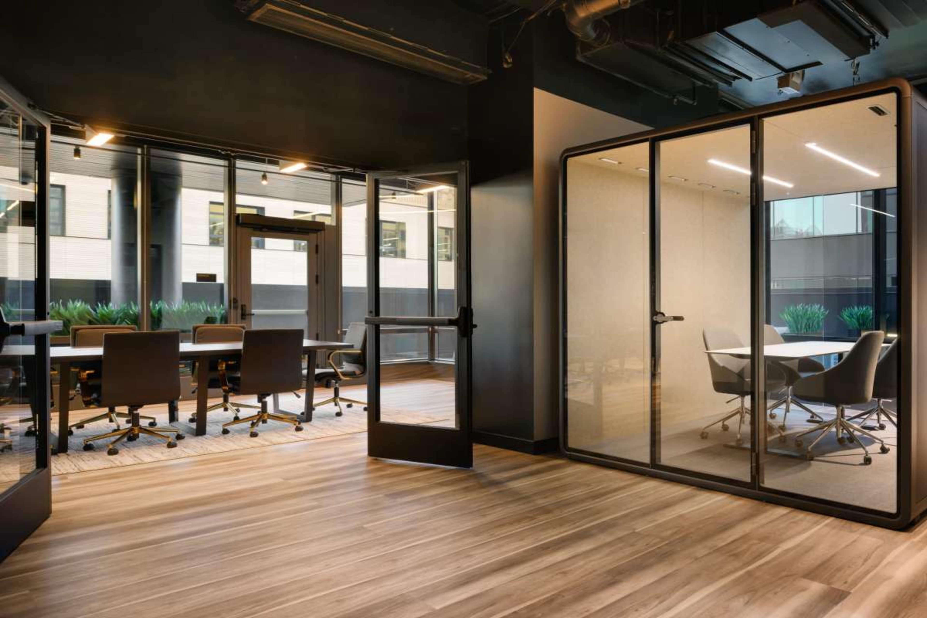 The image shows a modern office space featuring a glass-walled conference room and a workspace with wooden flooring and contemporary furniture.