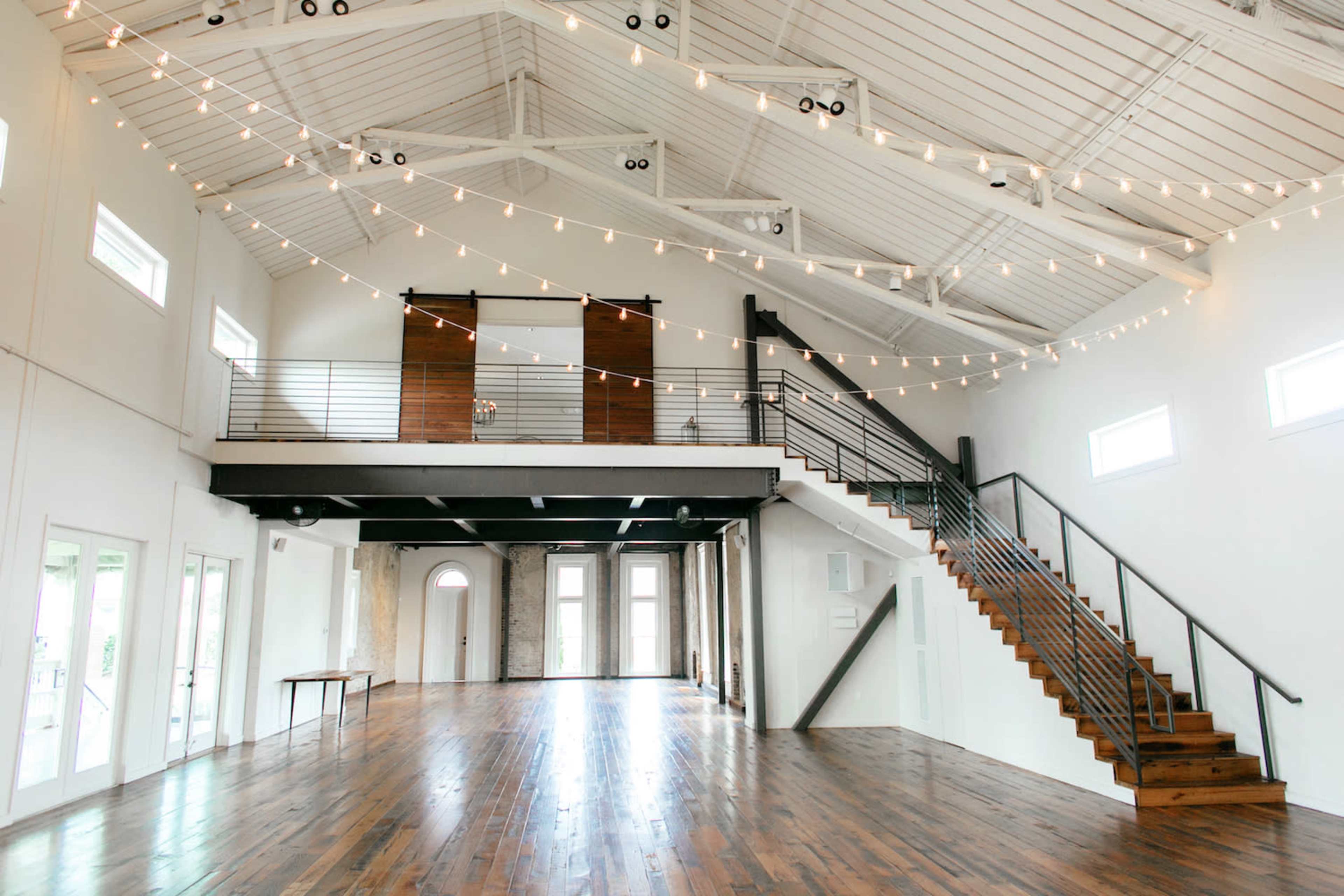 A spacious indoor venue with high ceilings, wooden floors, and string lights, featuring a staircase and large windows.