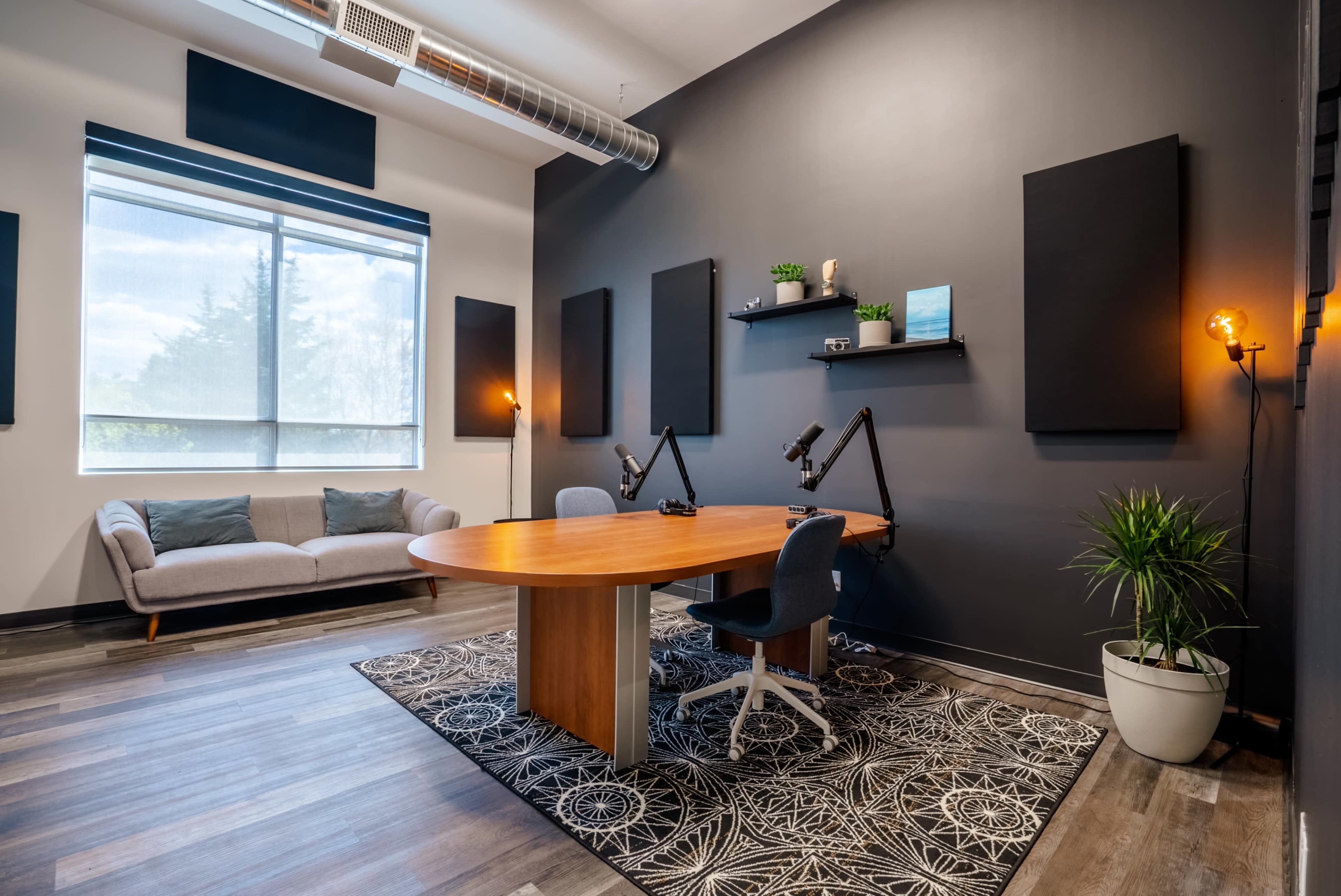 A modern office space features a round wooden table with two adjustable desk microphones, a gray sofa, plants, and black acoustic wall panels against a dark wall.