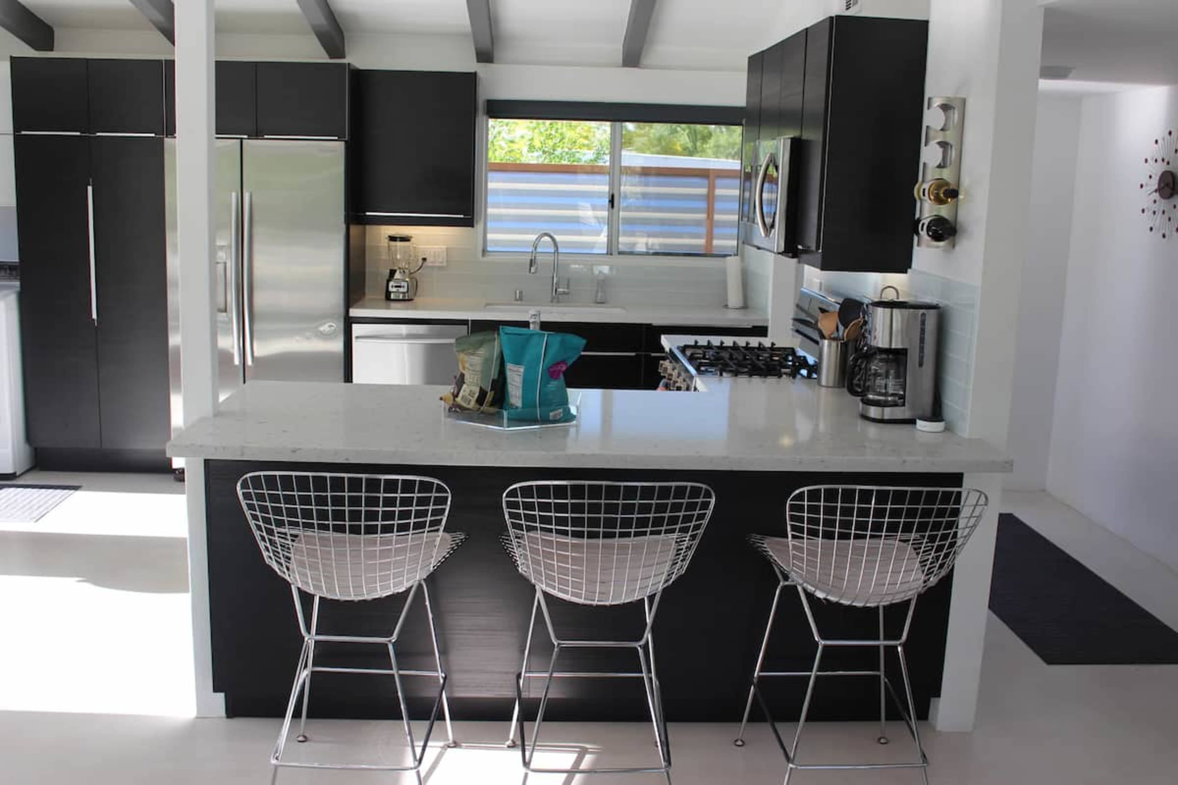 The image shows a modern kitchen with black cabinetry, stainless steel appliances, a gray countertop, and three wire bar stools arranged around a kitchen island.