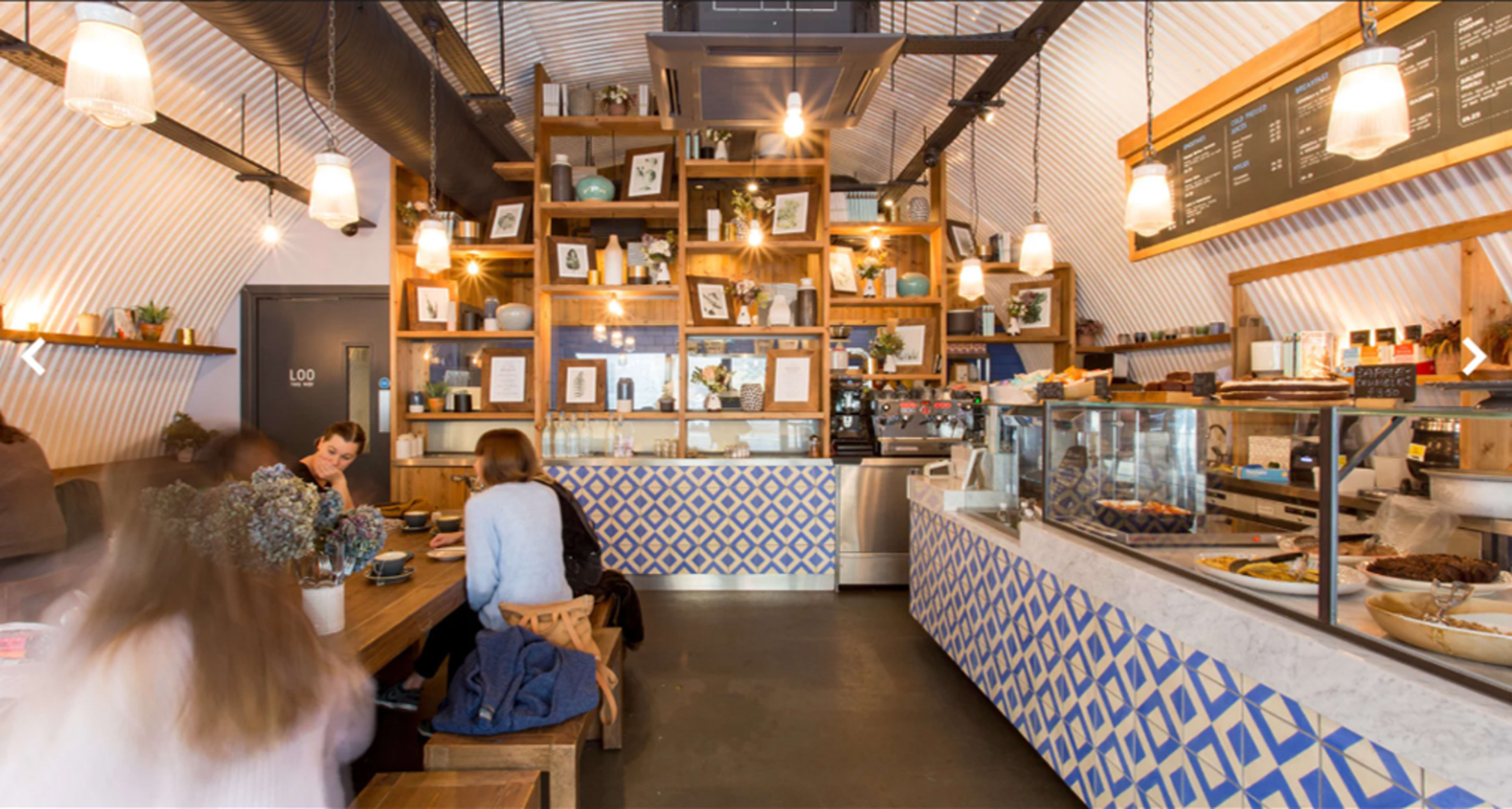 A cozy café interior features wooden shelves filled with plants and photos, a marble counter displaying pastries, and patrons seated at tables.