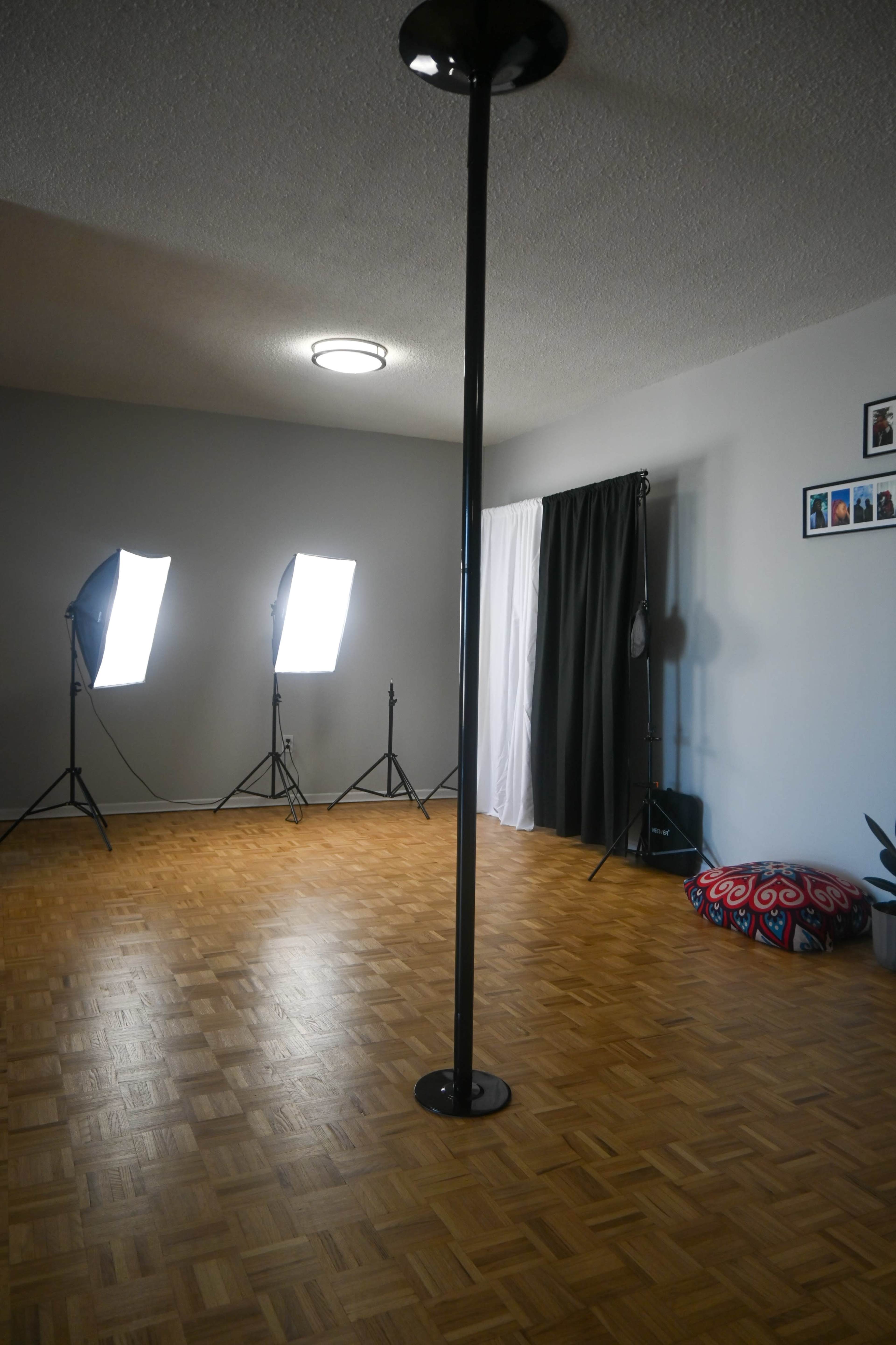 A spacious room features a central pole, two bright studio lights on tripods, a curtain, and several decorative items on the walls.