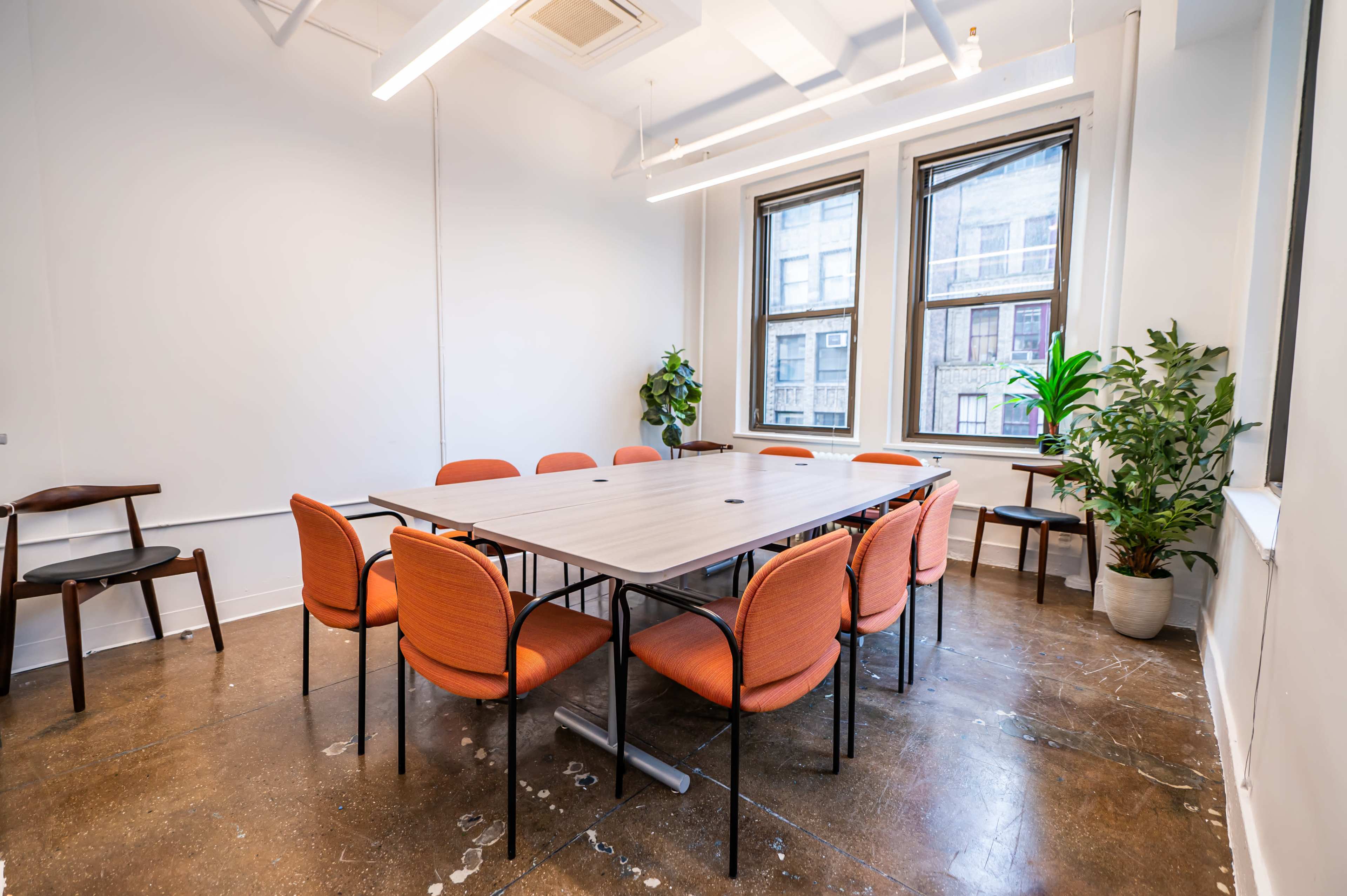 A conference room features a large rectangular table surrounded by eight orange chairs, with windows allowing natural light and plants in the corners.