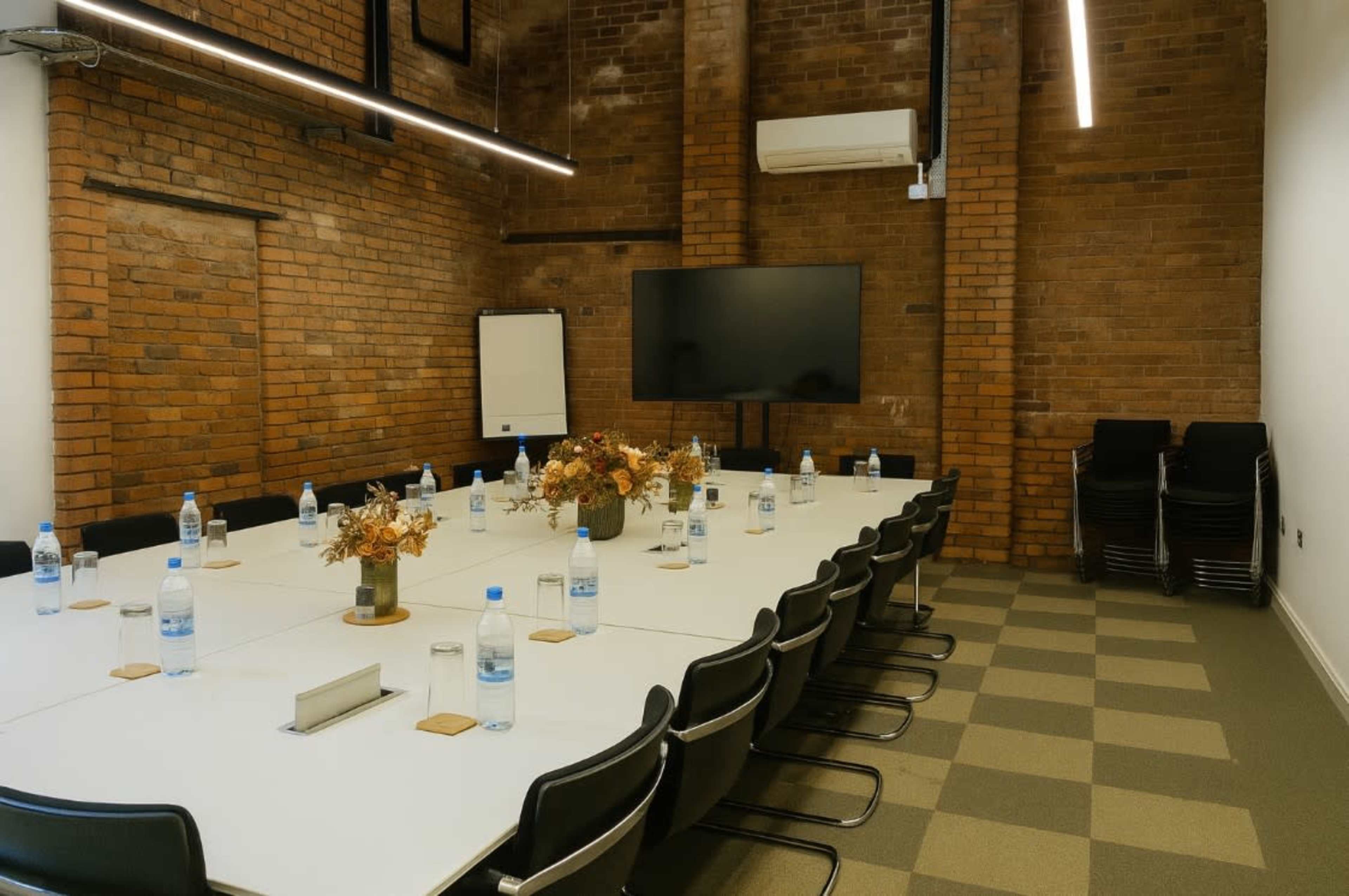 A conference room features a large table surrounded by chairs, with a screen mounted on the wall and water bottles placed on the table.