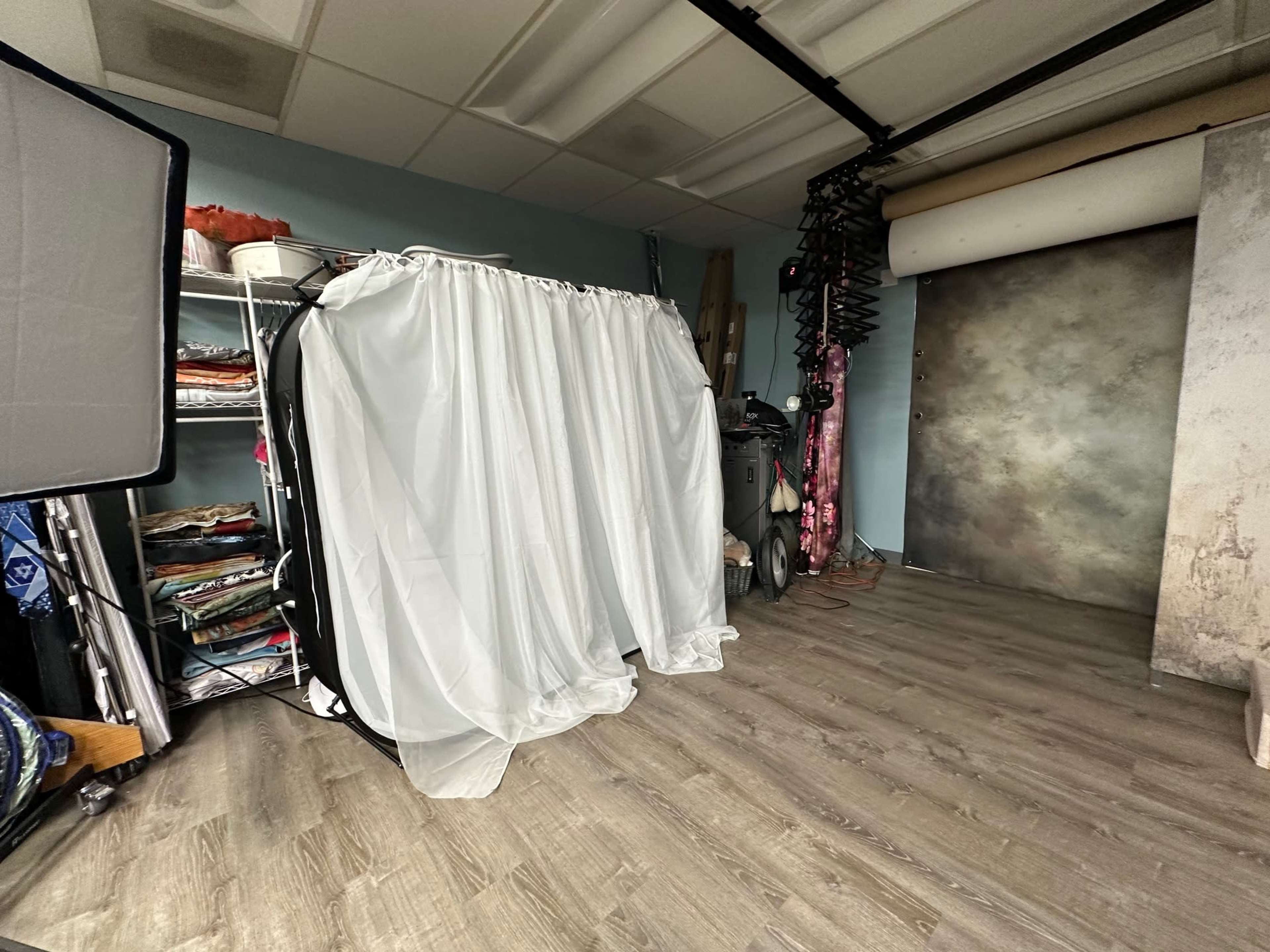 The image shows a photography studio with a white curtain in front of a shelving unit and a backdrop on one side.