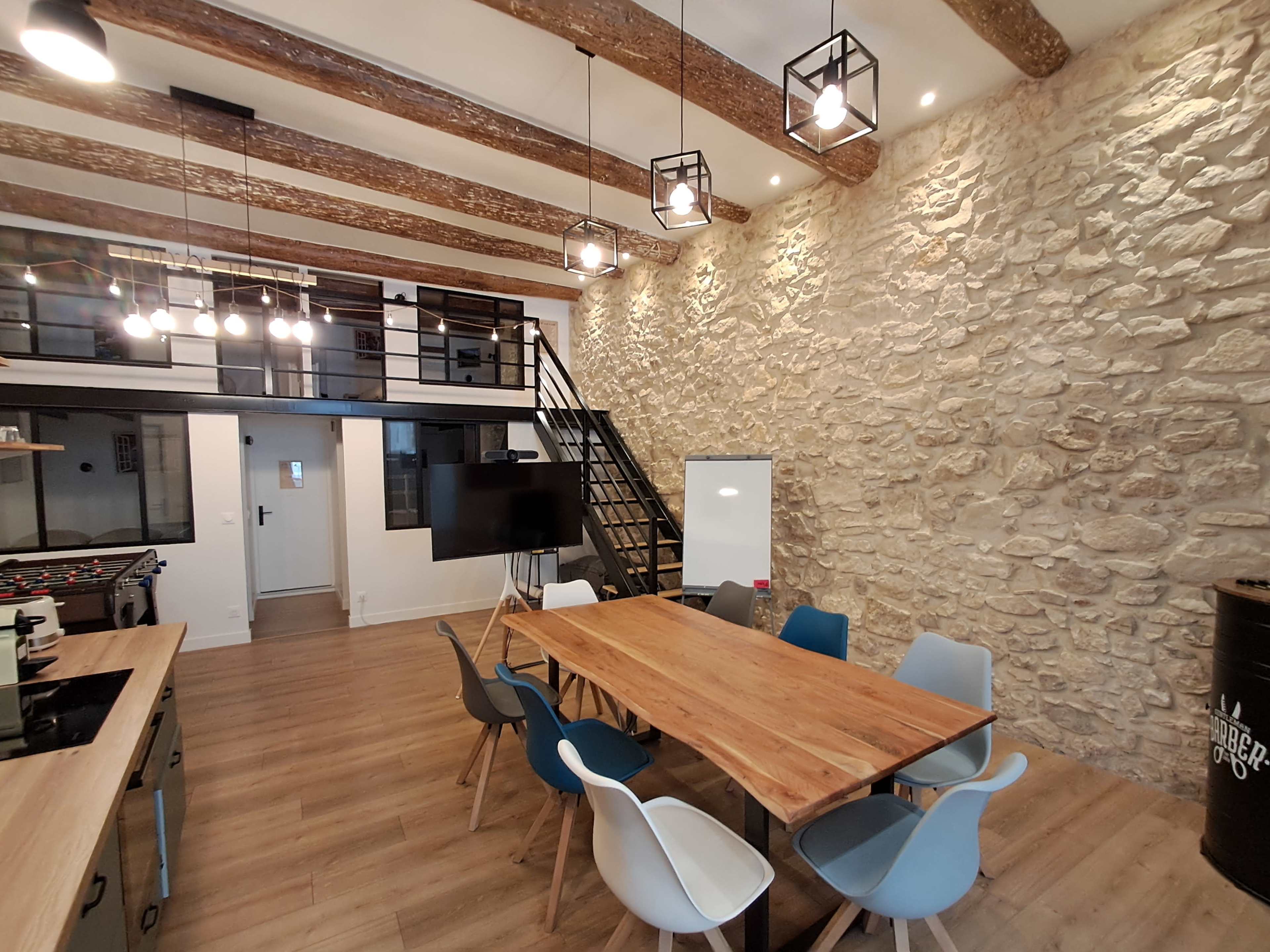 The image shows a modern meeting room with a wooden table surrounded by chairs, a stone wall, and a staircase leading to a loft area.