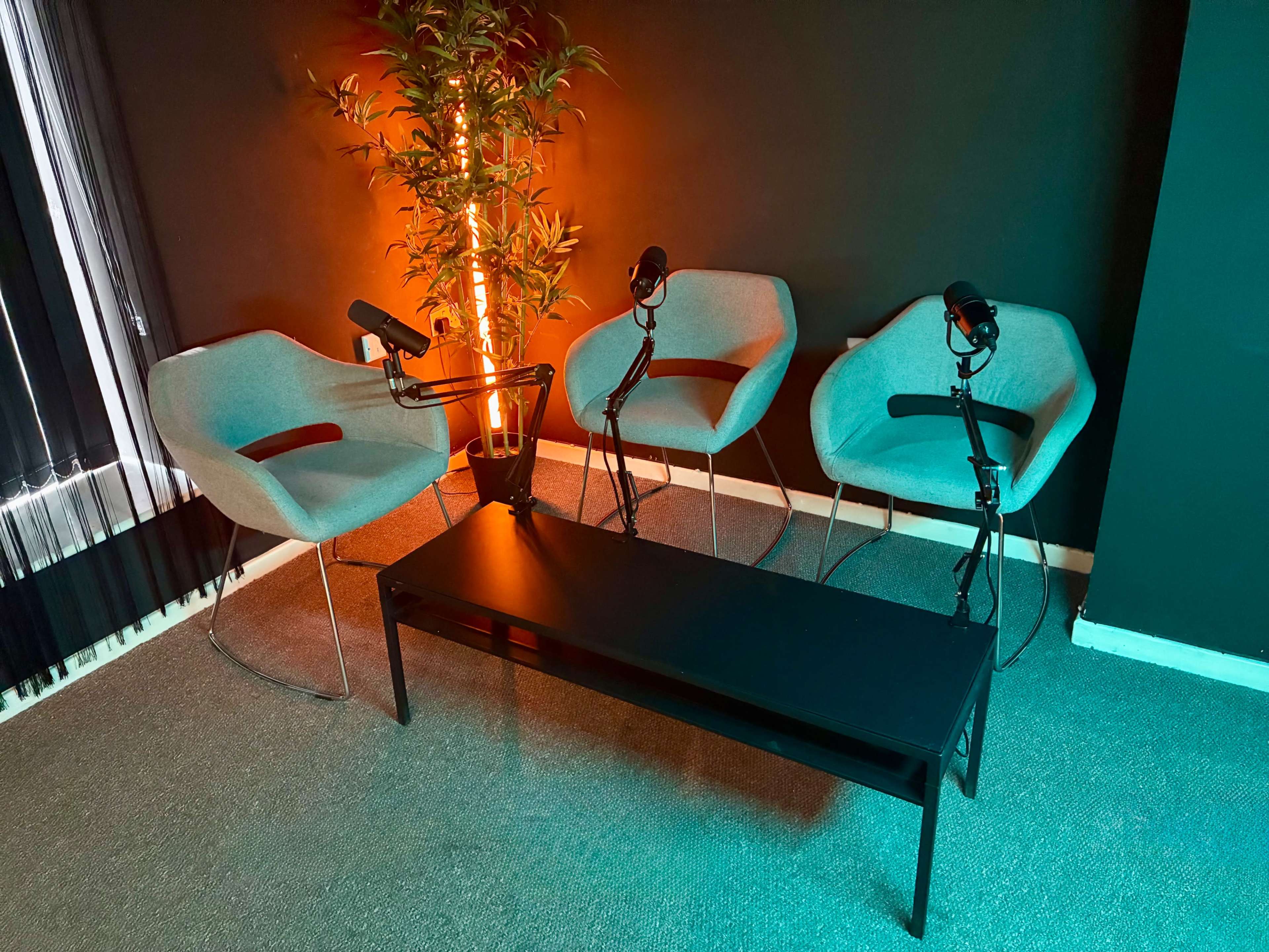 The image shows a small studio setup with three gray chairs and a black table, accompanied by microphones and an illuminated plant against a dark wall.