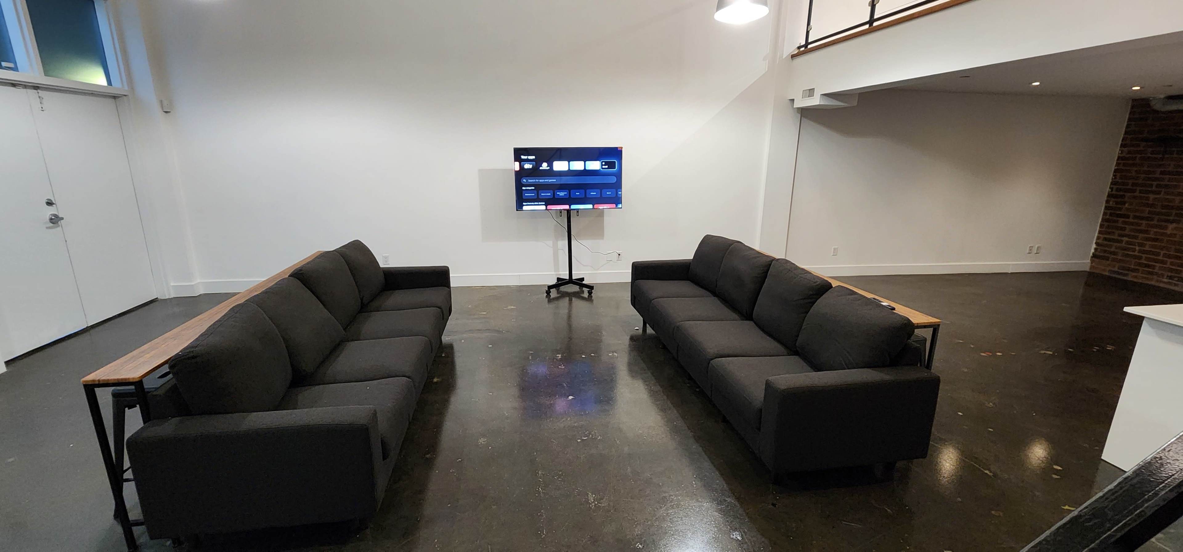 Two black sofas facing a mounted television in a spacious room with a polished concrete floor and white walls.
