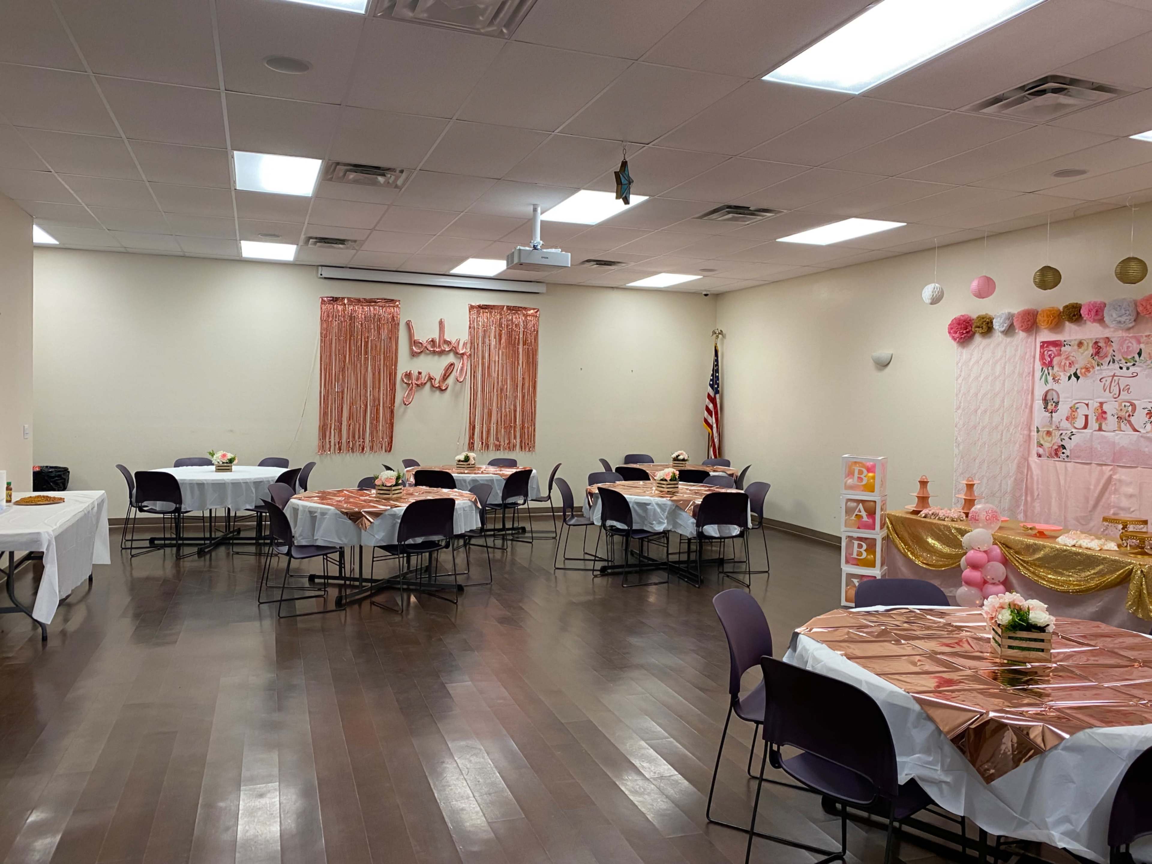 The image shows a decorated event space with several round tables covered in white tablecloths, pink and gold accents, and a backdrop reading "baby girl."