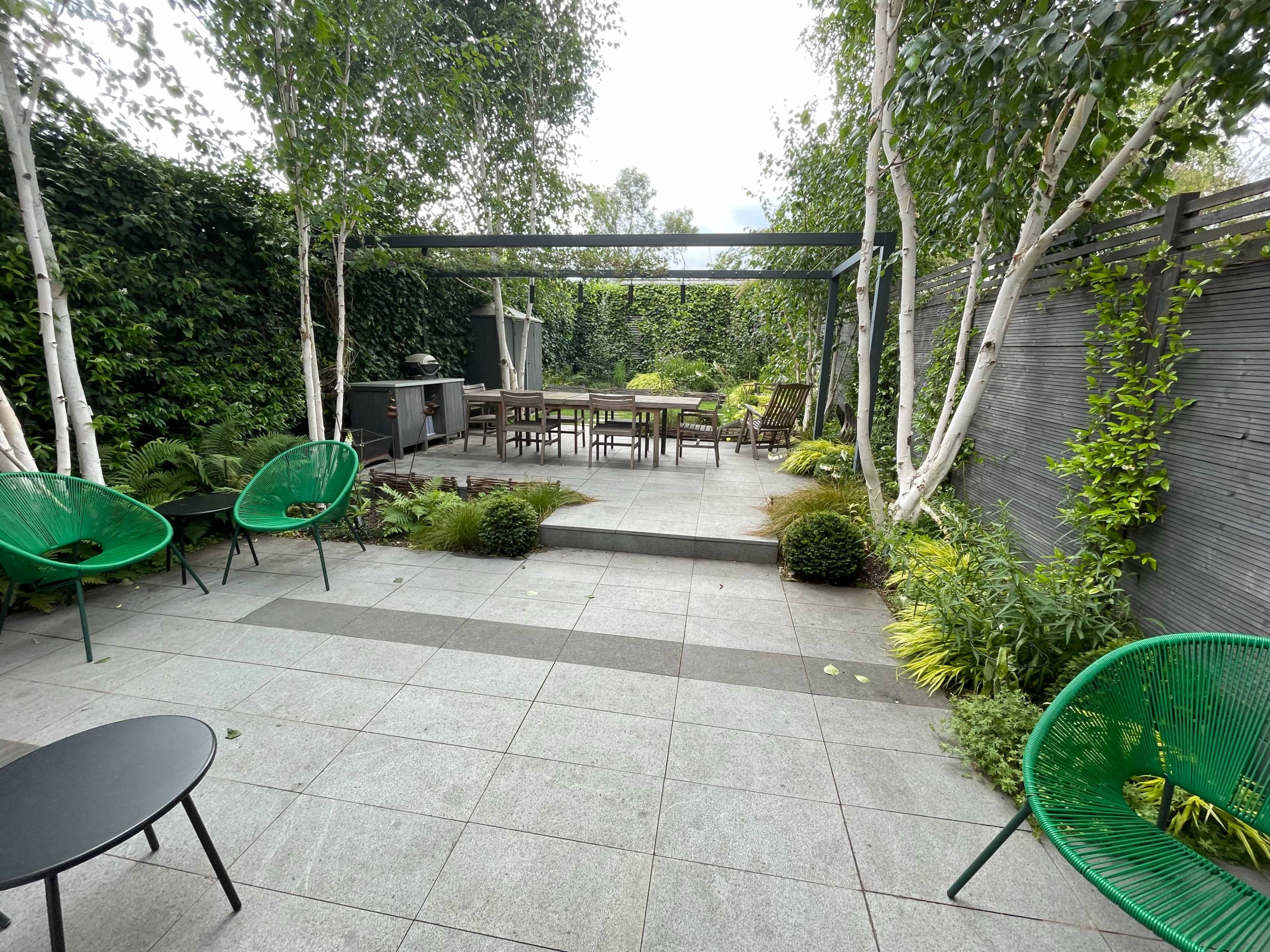 A modern garden features green chairs and a dining table surrounded by lush plants and trees.