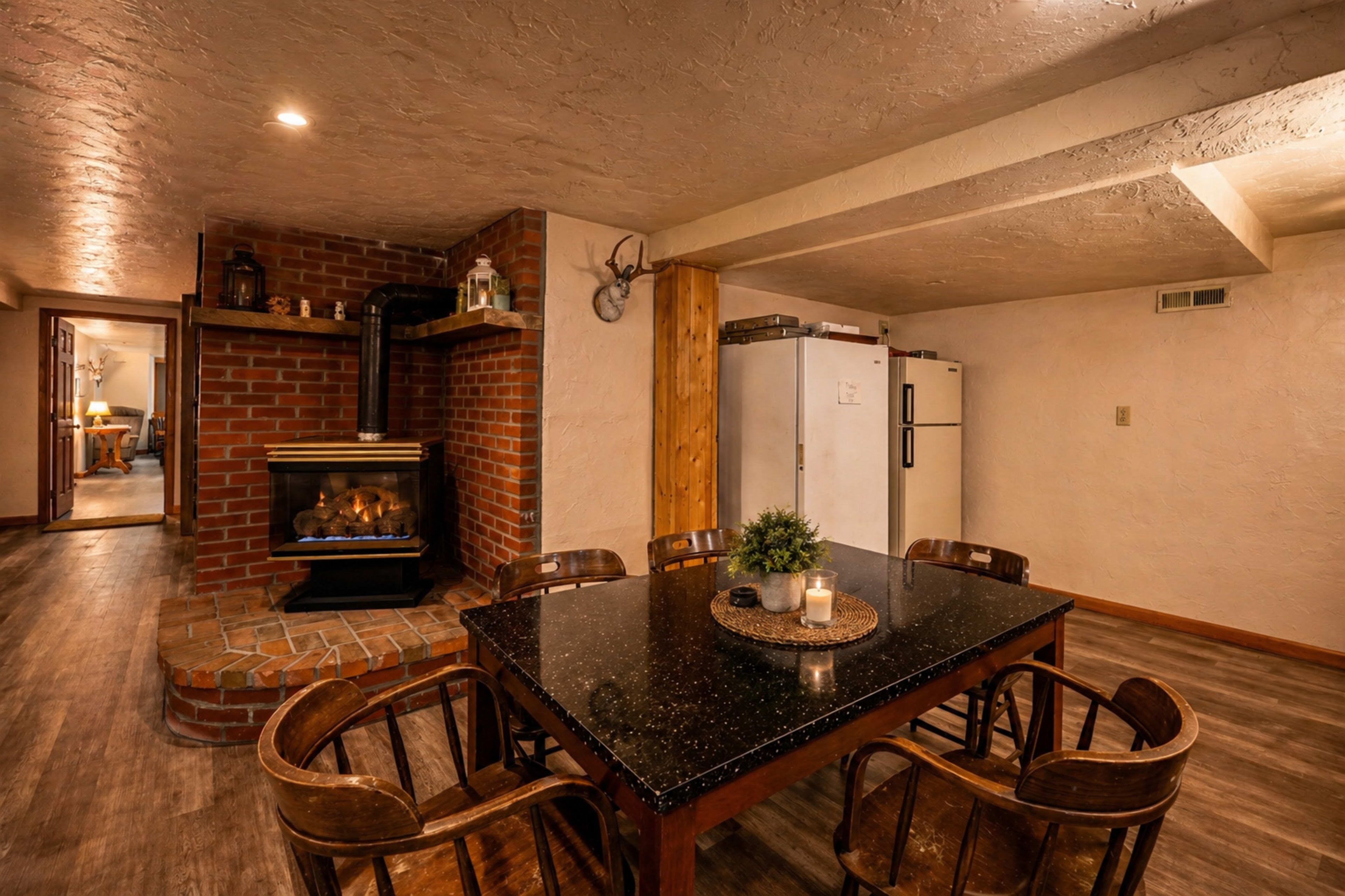 A dining area features a dark granite table surrounded by wooden chairs, with a brick fireplace and a white refrigerator in the background.