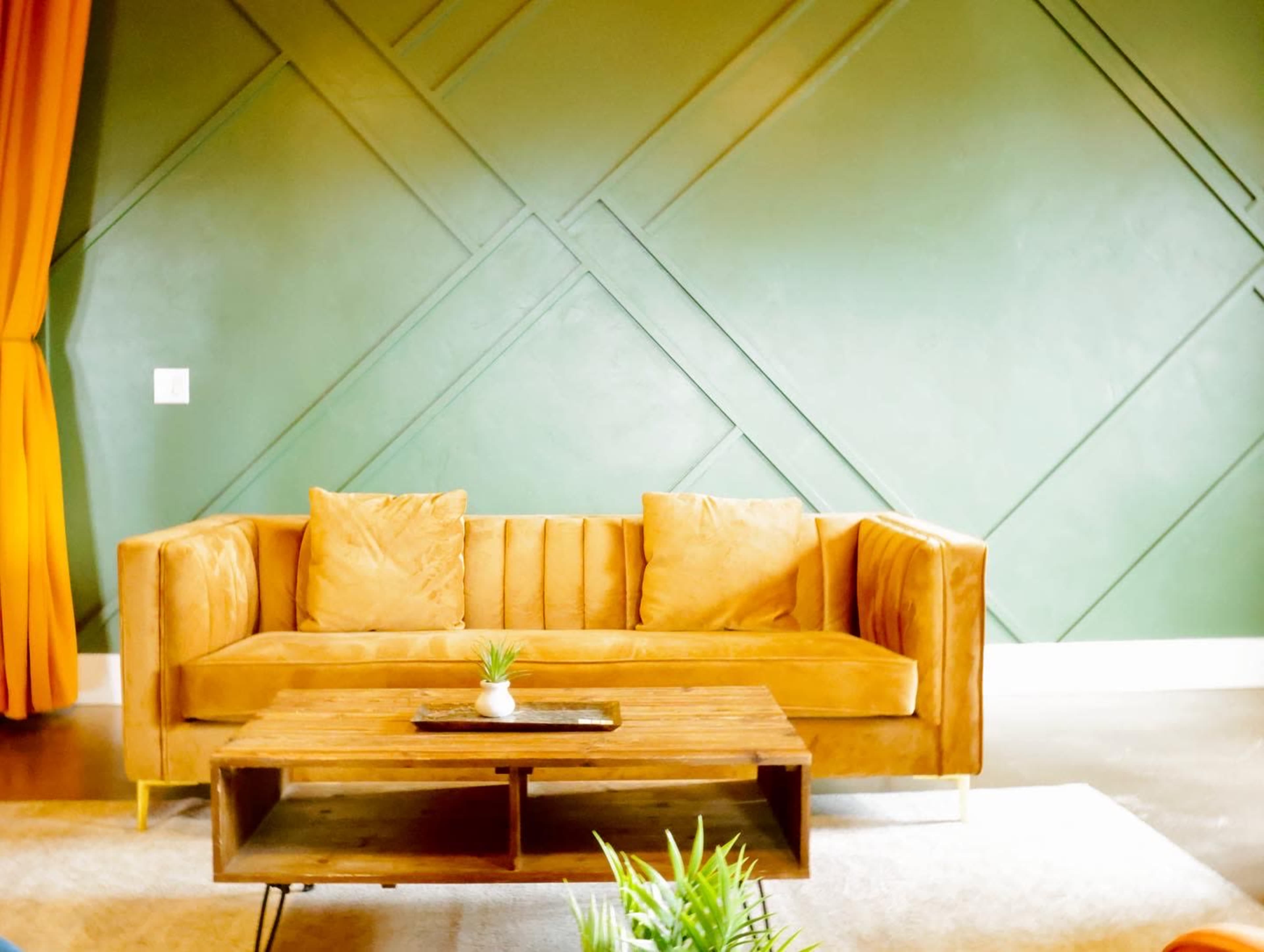 A mustard-colored couch with two cushions in front of a wooden coffee table, all set against a green geometric-patterned wall and orange curtains.