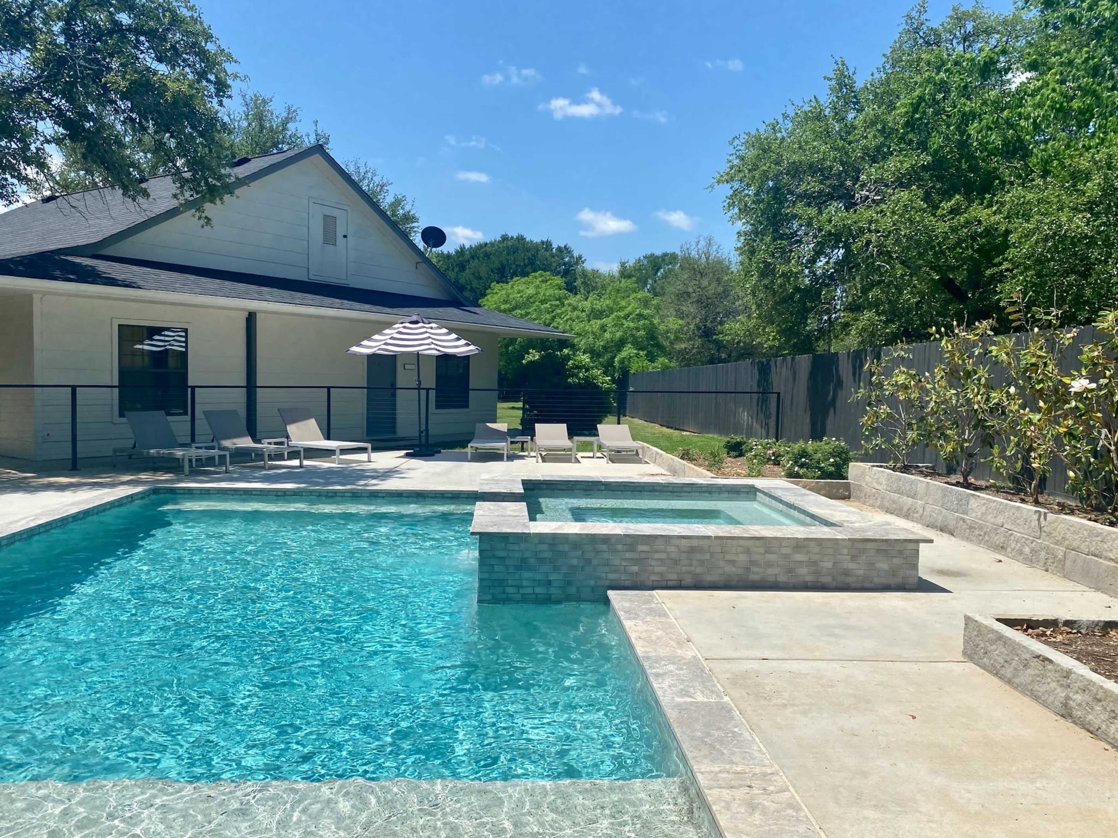A backyard features a swimming pool and a spa, surrounded by lounge chairs and shaded by a striped umbrella, with a house and greenery in the background.