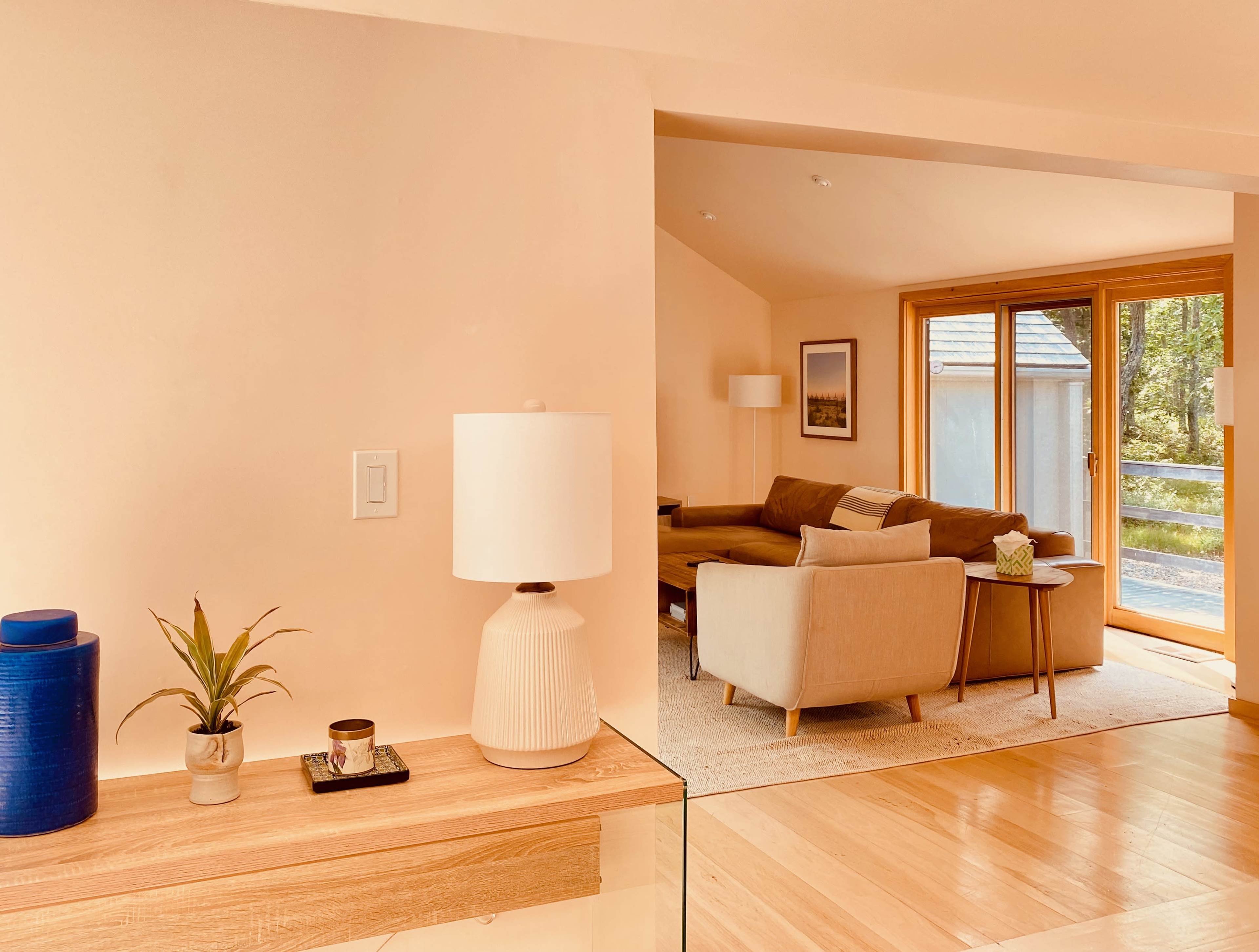 A cozy living space features a beige color palette with modern furniture, a lamp, and a plant, visible through a doorway.