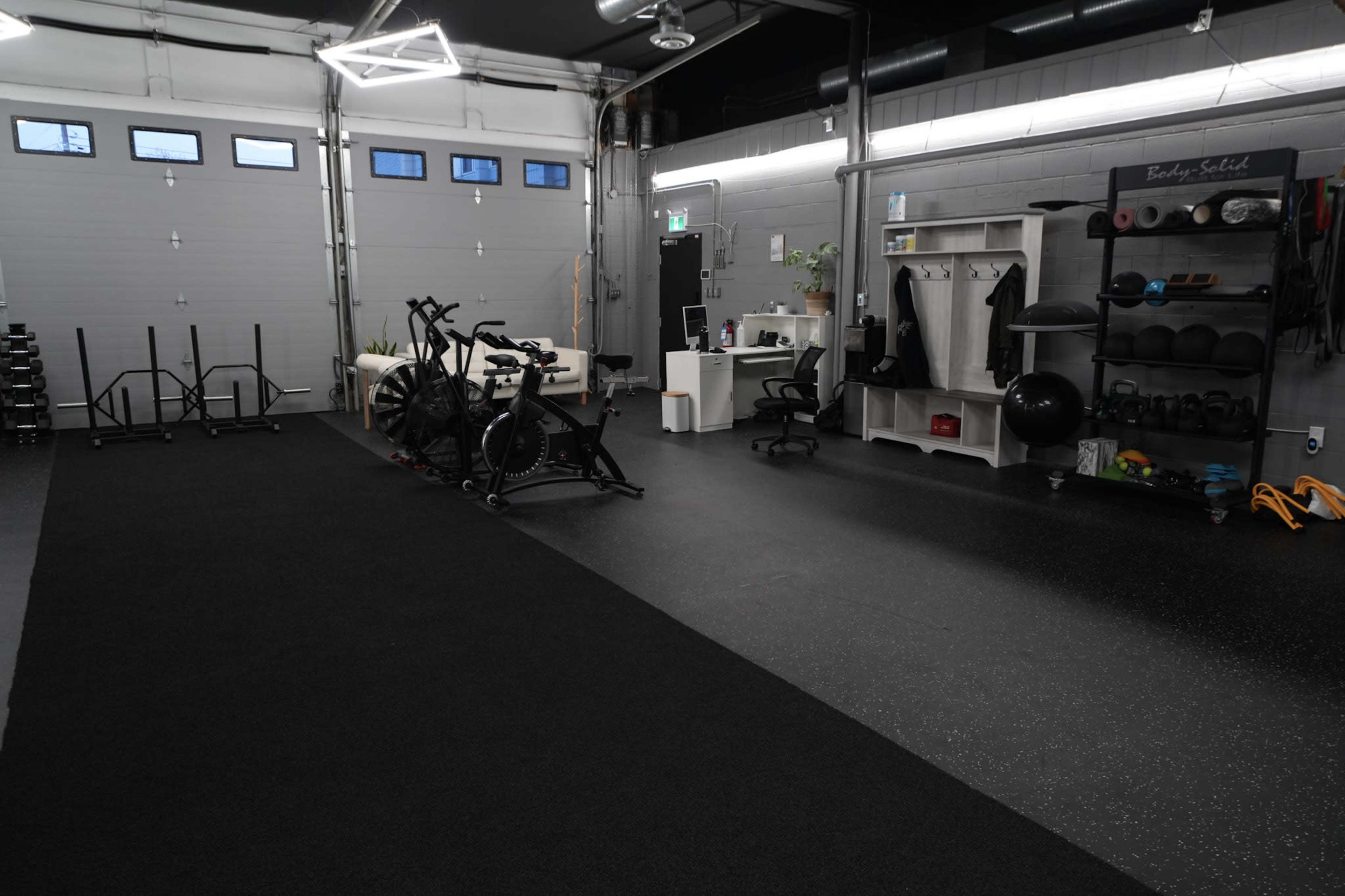 The image shows a spacious gym with exercise equipment on one side, including bikes and weights, and an area with a desk and organization for workout gear.