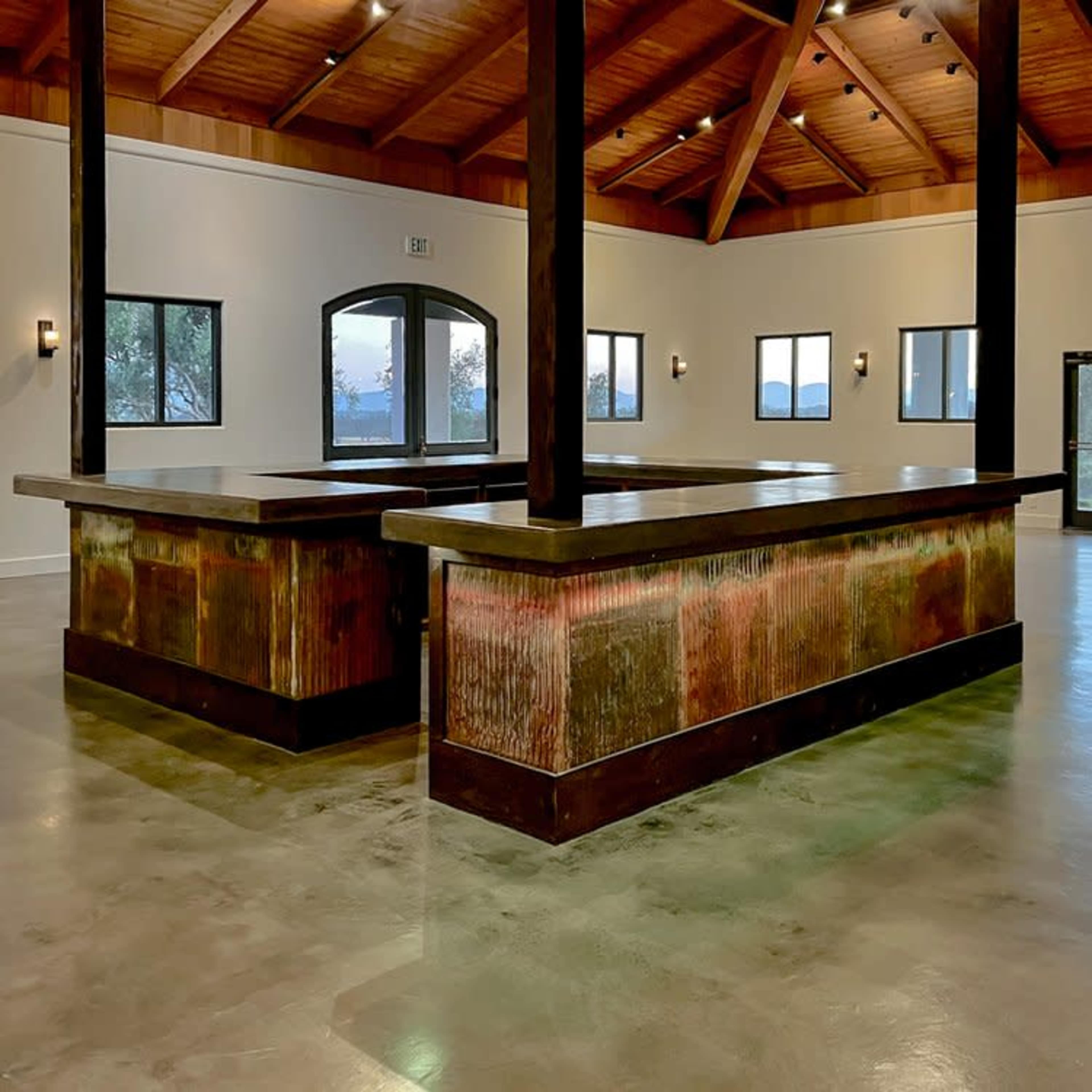 A spacious interior with two wooden bars, featuring a corrugated metal front, under a high ceiling with exposed beams and large windows.