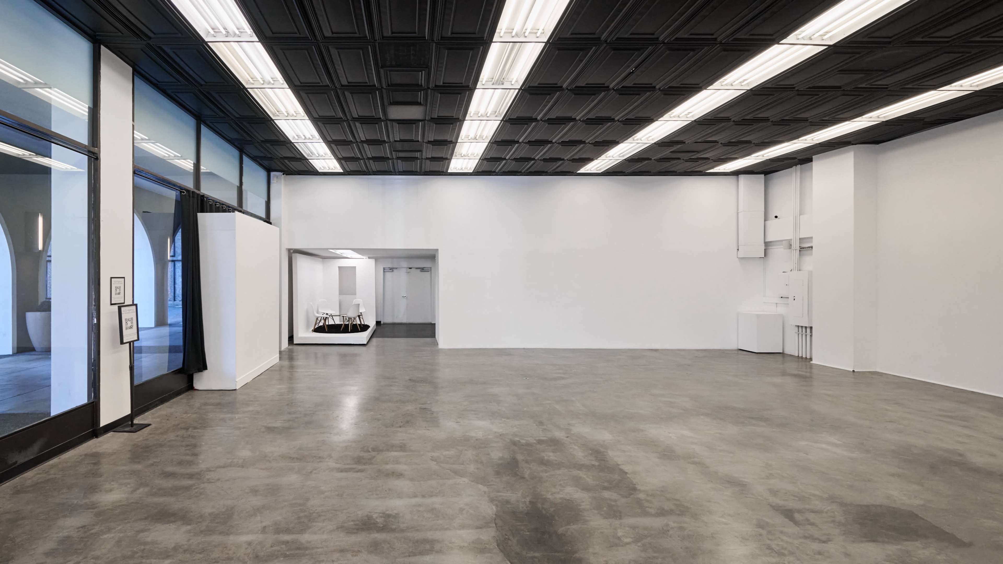 The image shows a spacious, empty interior with polished concrete flooring, white walls, and a grid-patterned black ceiling.