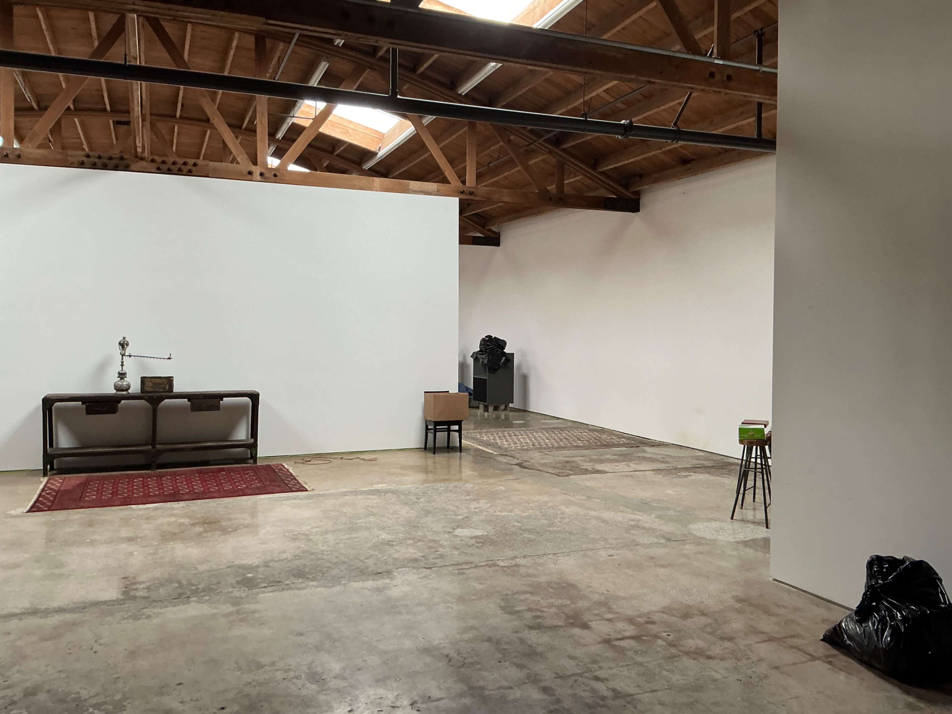 The image shows an empty, spacious room with wooden beams, a red rug, a table and chair, and a waste bag in the corner.