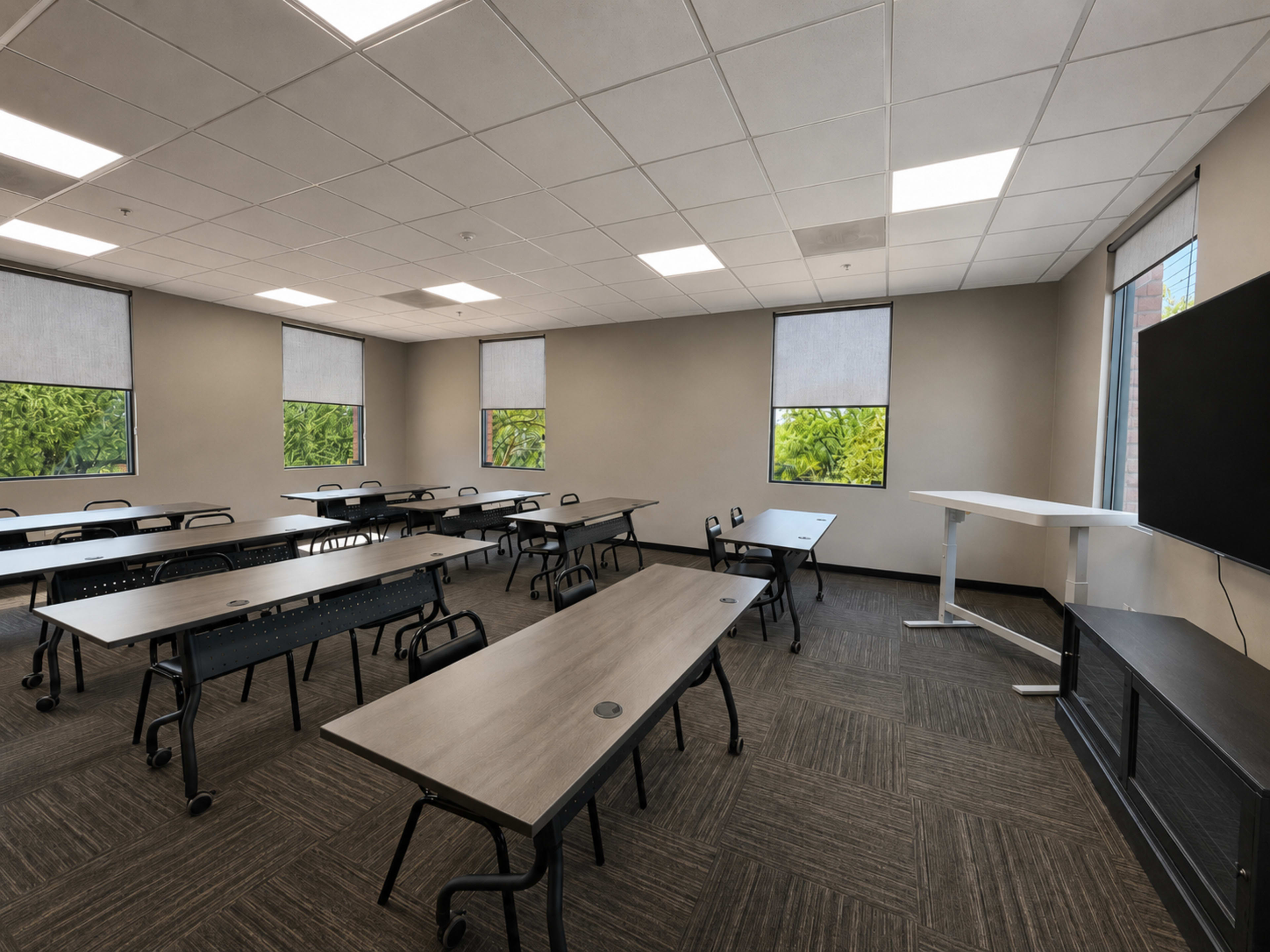 The image shows a spacious classroom filled with multiple rows of tables and chairs, featuring large windows with greenery visible outside.