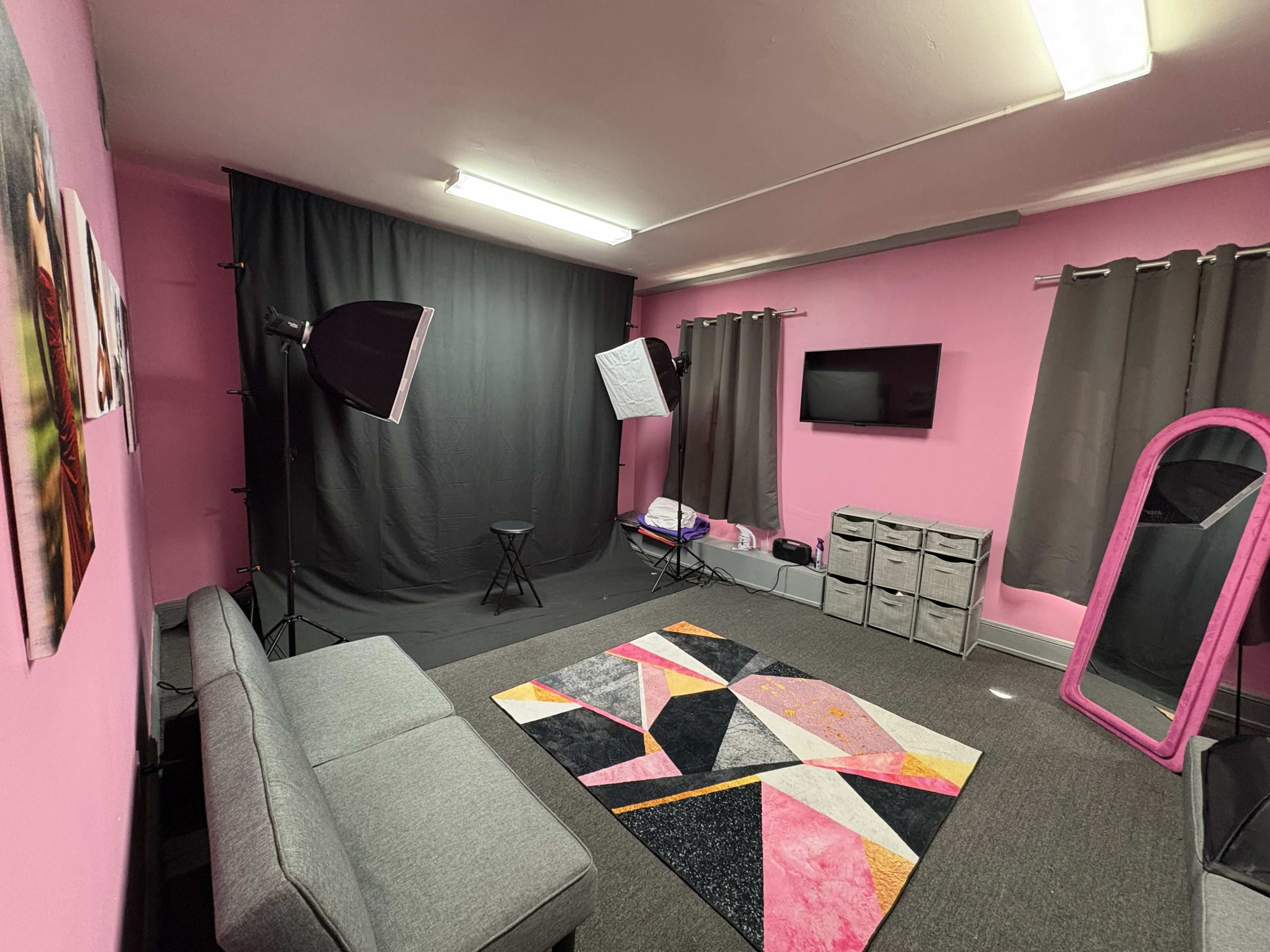 The image shows a small studio space with pink walls, gray curtains, a gray couch, a patterned rug, and photography lighting equipment.
