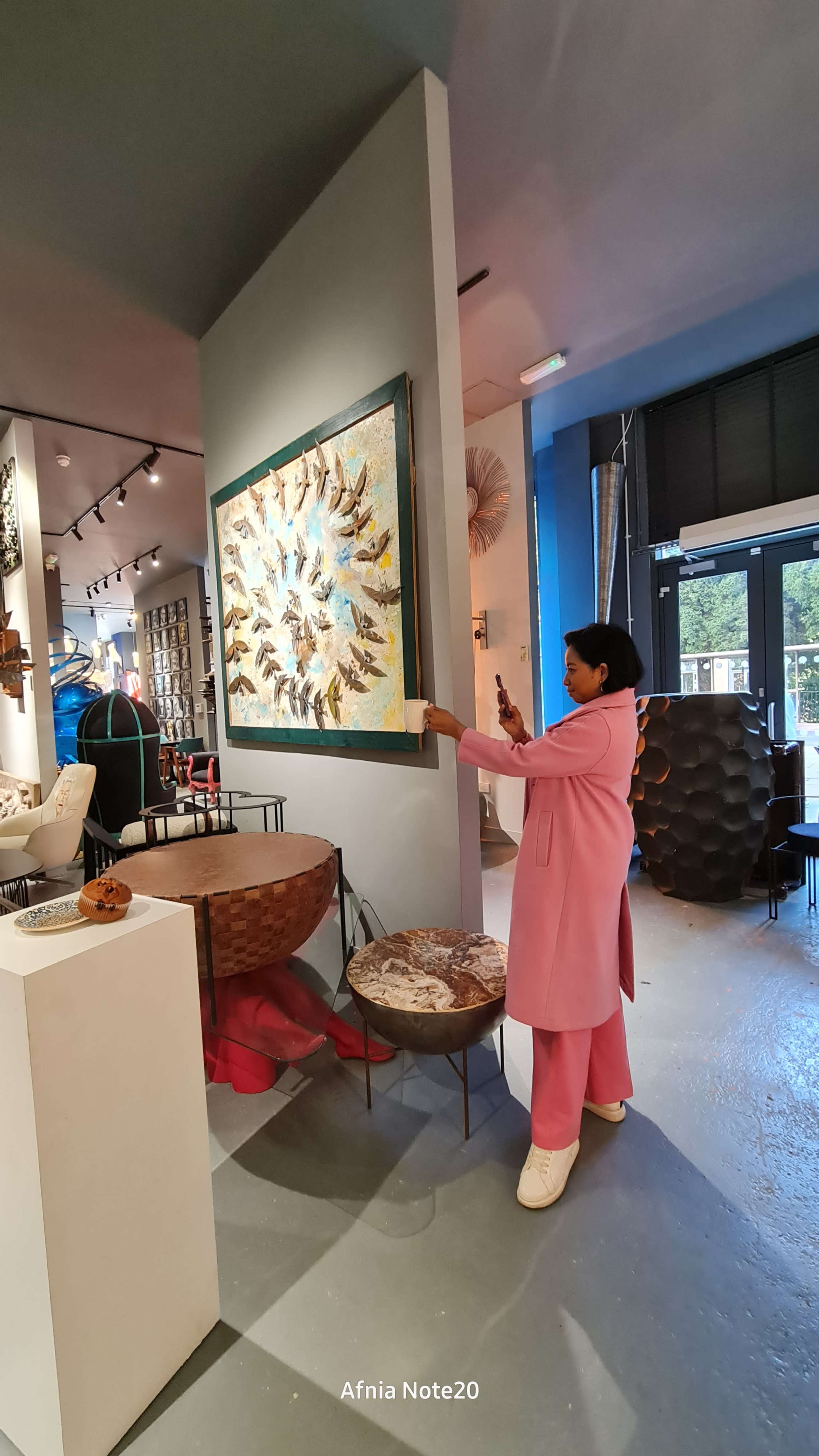 A woman in a pink outfit is standing in an art gallery, adjusting a large framed artwork on the wall.