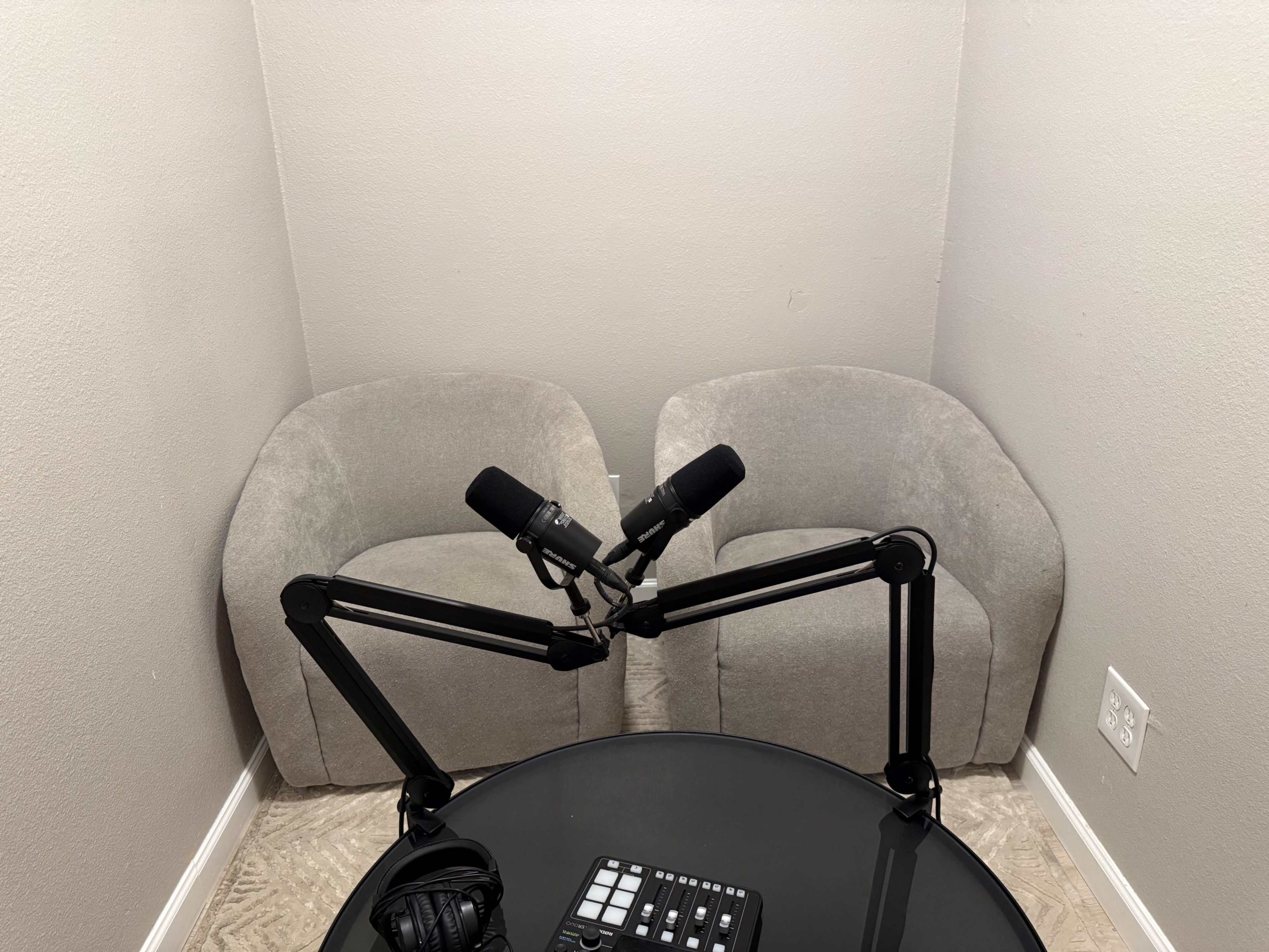 A small, sparsely furnished recording studio features two gray chairs positioned opposite each other with microphones mounted on adjustable arms above a black table.