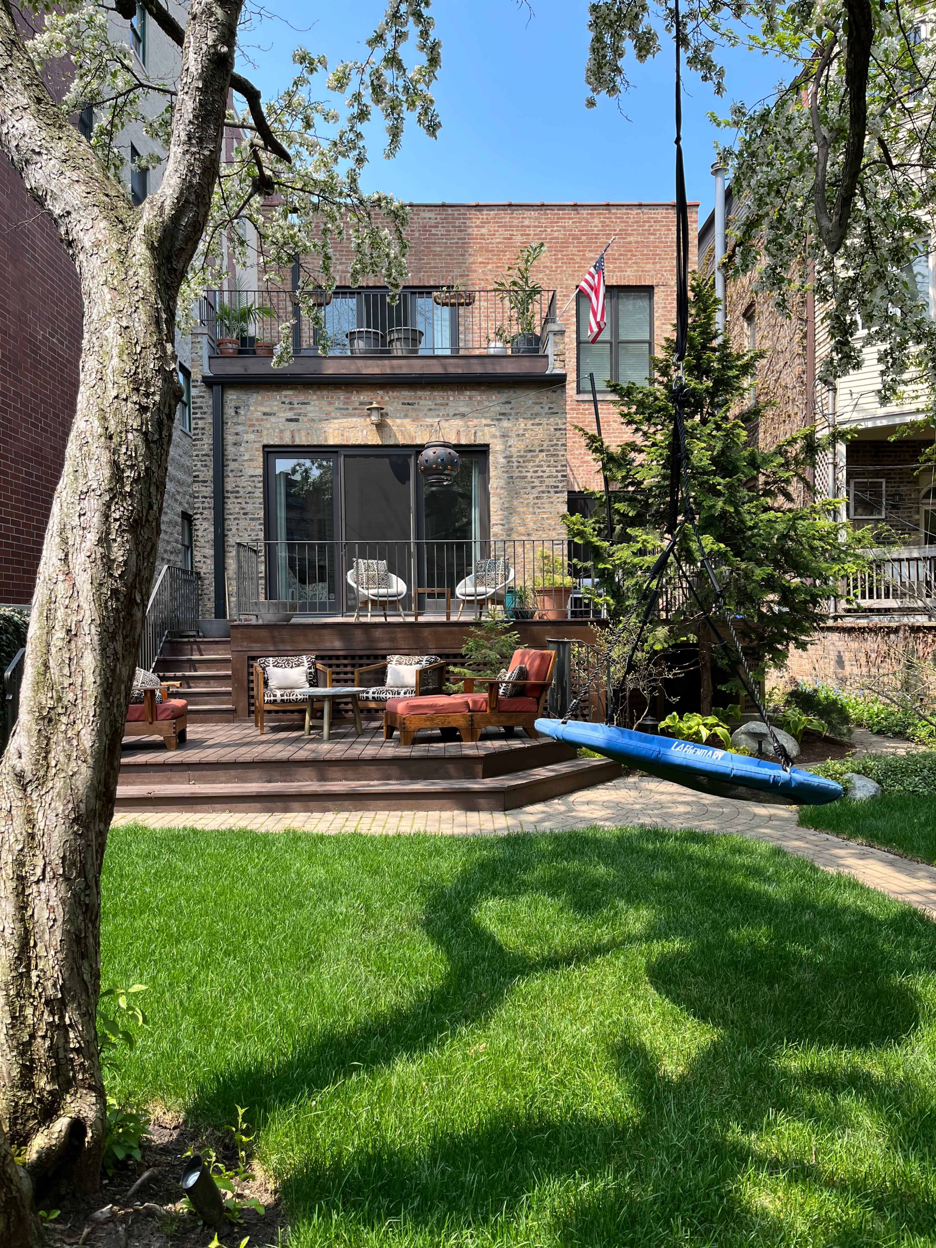 A backyard features a wooden deck with seating and a blue swing hanging from a tree, with a brick building and an American flag in the background.