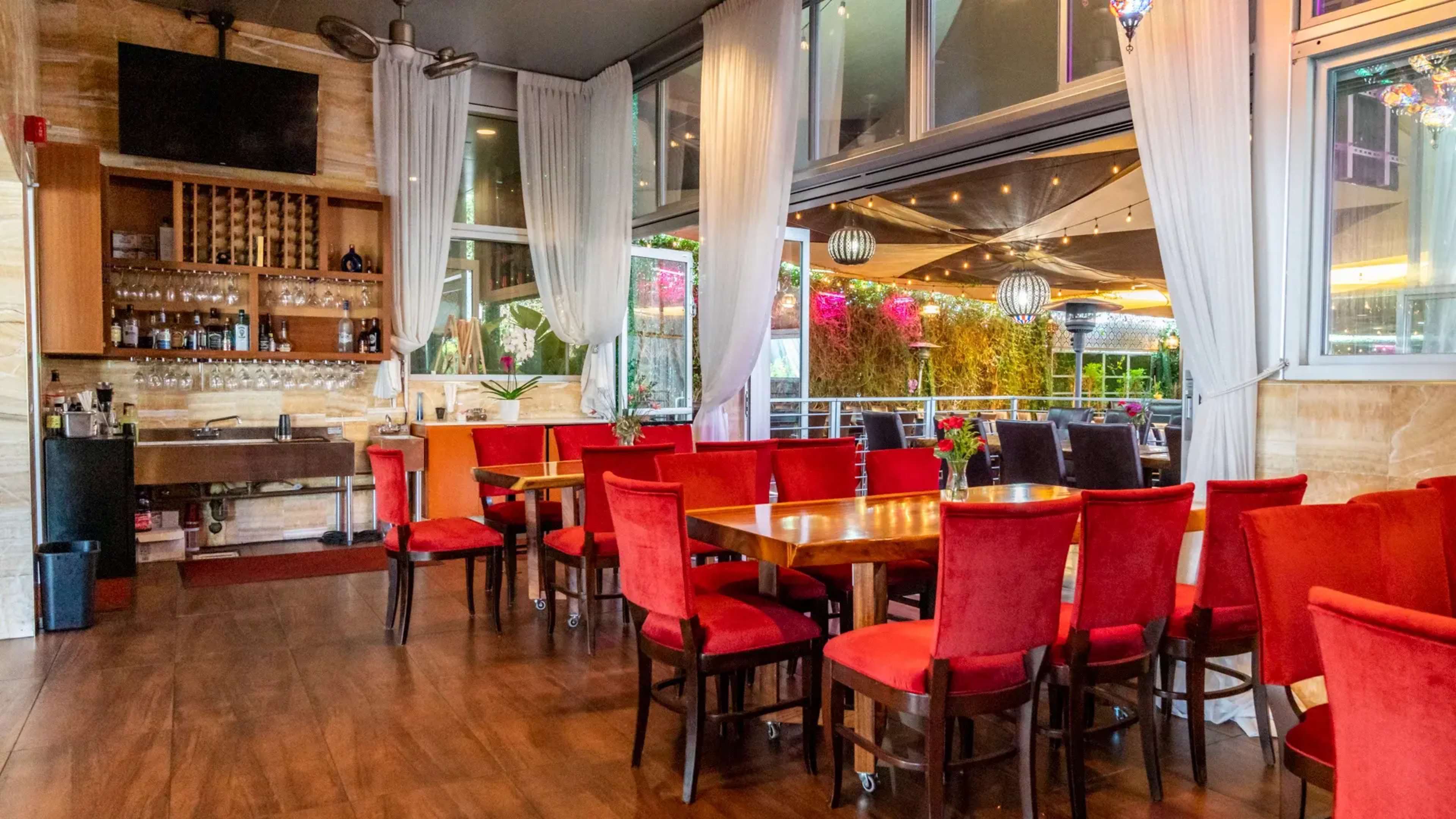 A restaurant interior with red upholstered chairs around wooden tables, large windows, and a bar area featuring shelves of bottles.