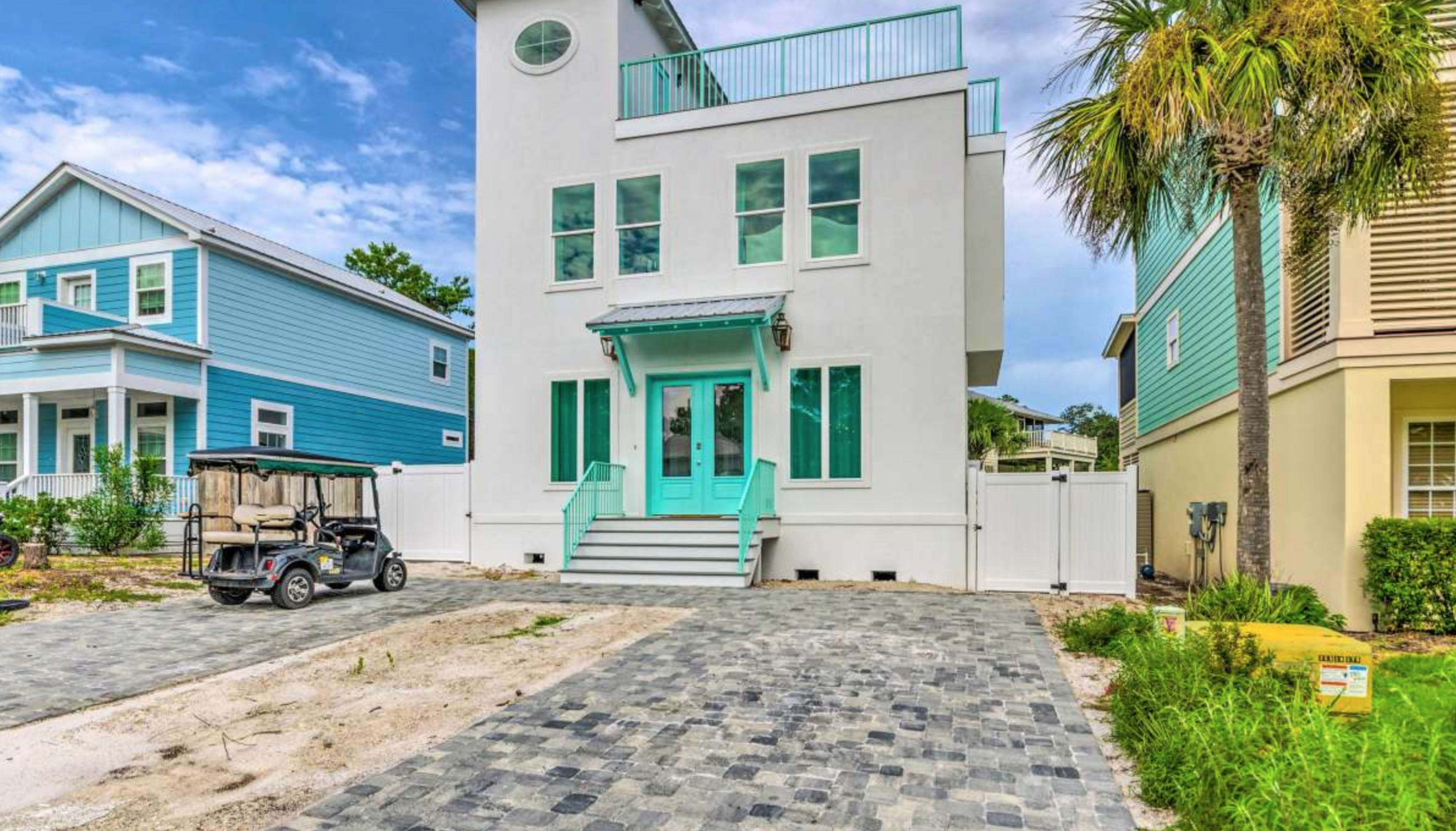 A modern two-story house with turquoise accents is situated at the end of a paved driveway, surrounded by other colorful homes and palm trees.