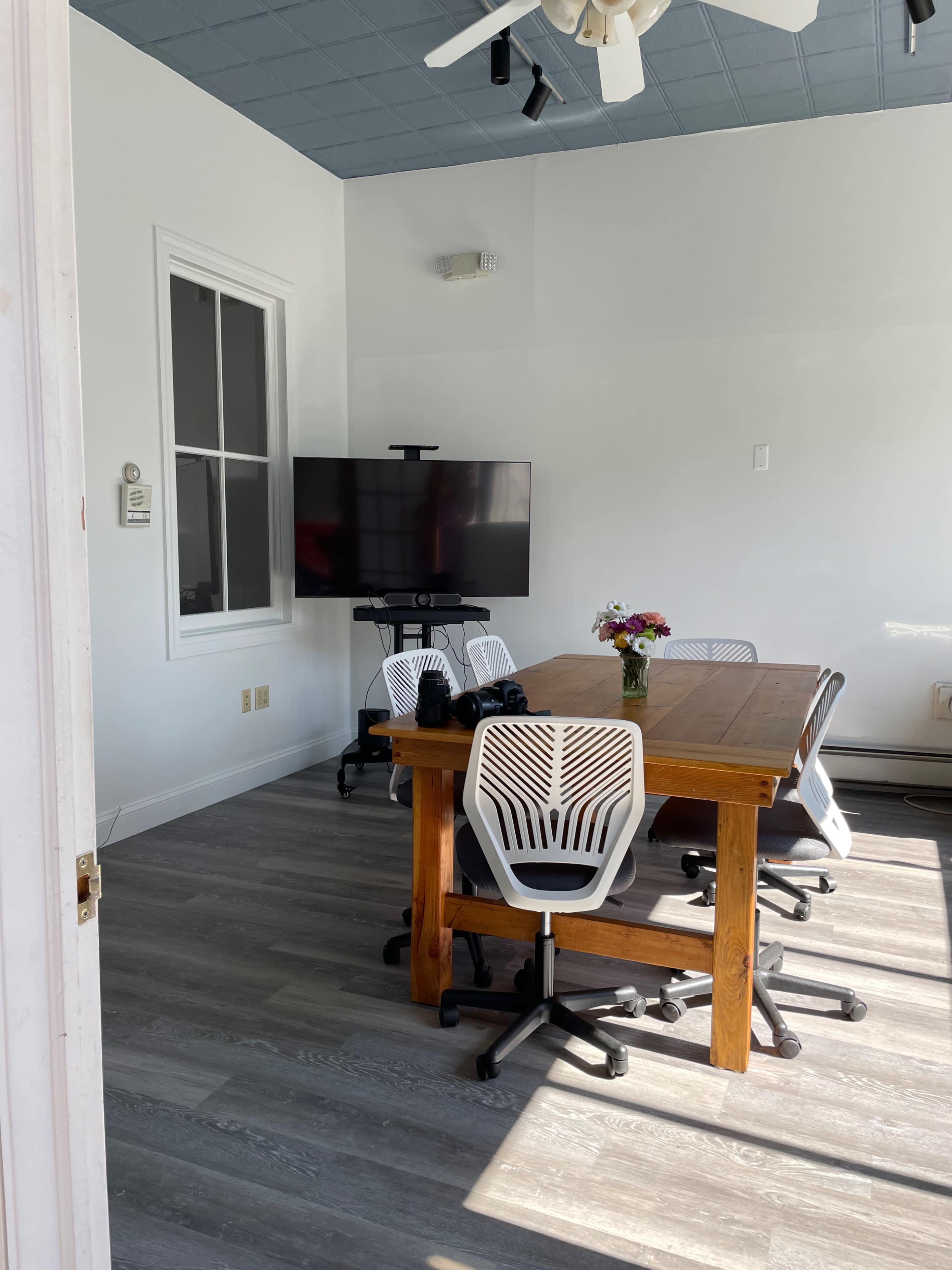 A conference room features a wooden table surrounded by six white chairs and a large television mounted on the wall.