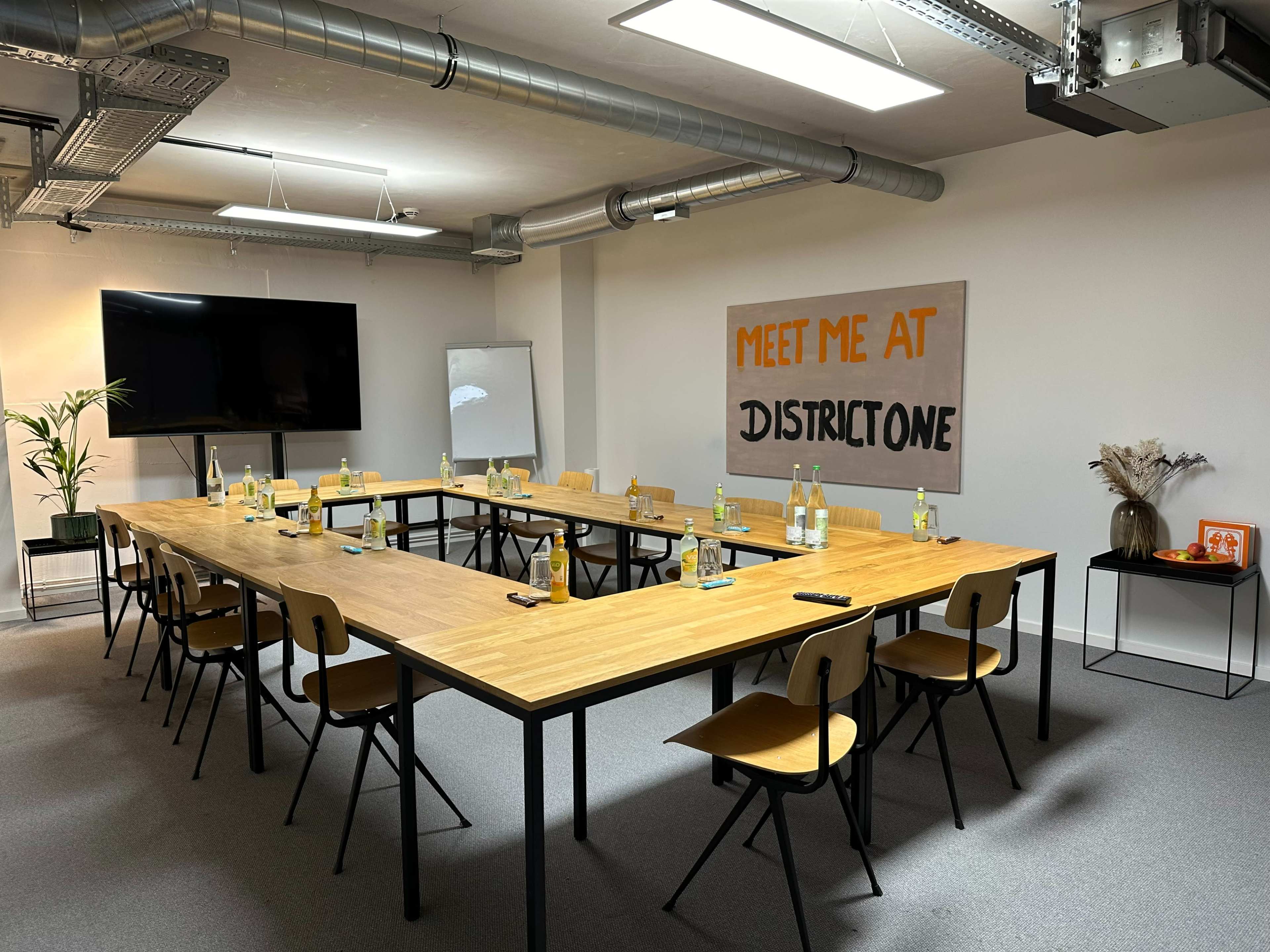 A conference room features a large rectangular table surrounded by chairs, with water bottles and glasses at each seat, and a sign reading "MEET ME AT DISTRICT ONE" displayed on the wall.