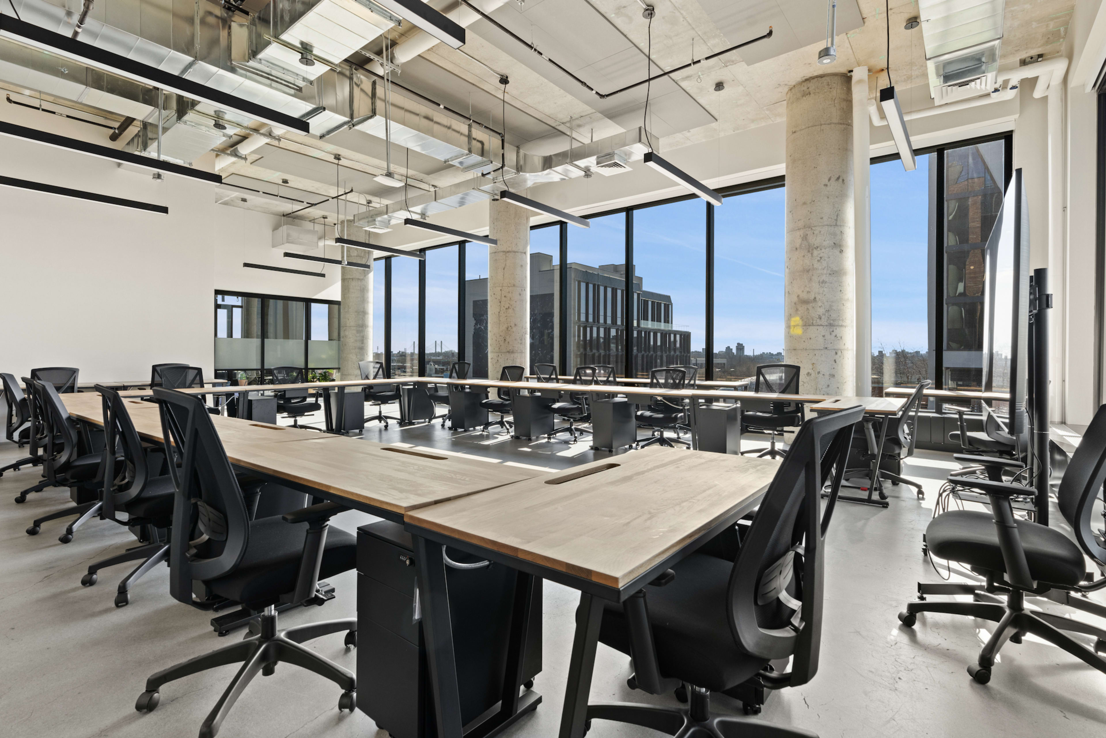 The image shows a modern office space with multiple large desks arranged in a U-shape, surrounded by black ergonomic chairs and large windows allowing plenty of natural light.