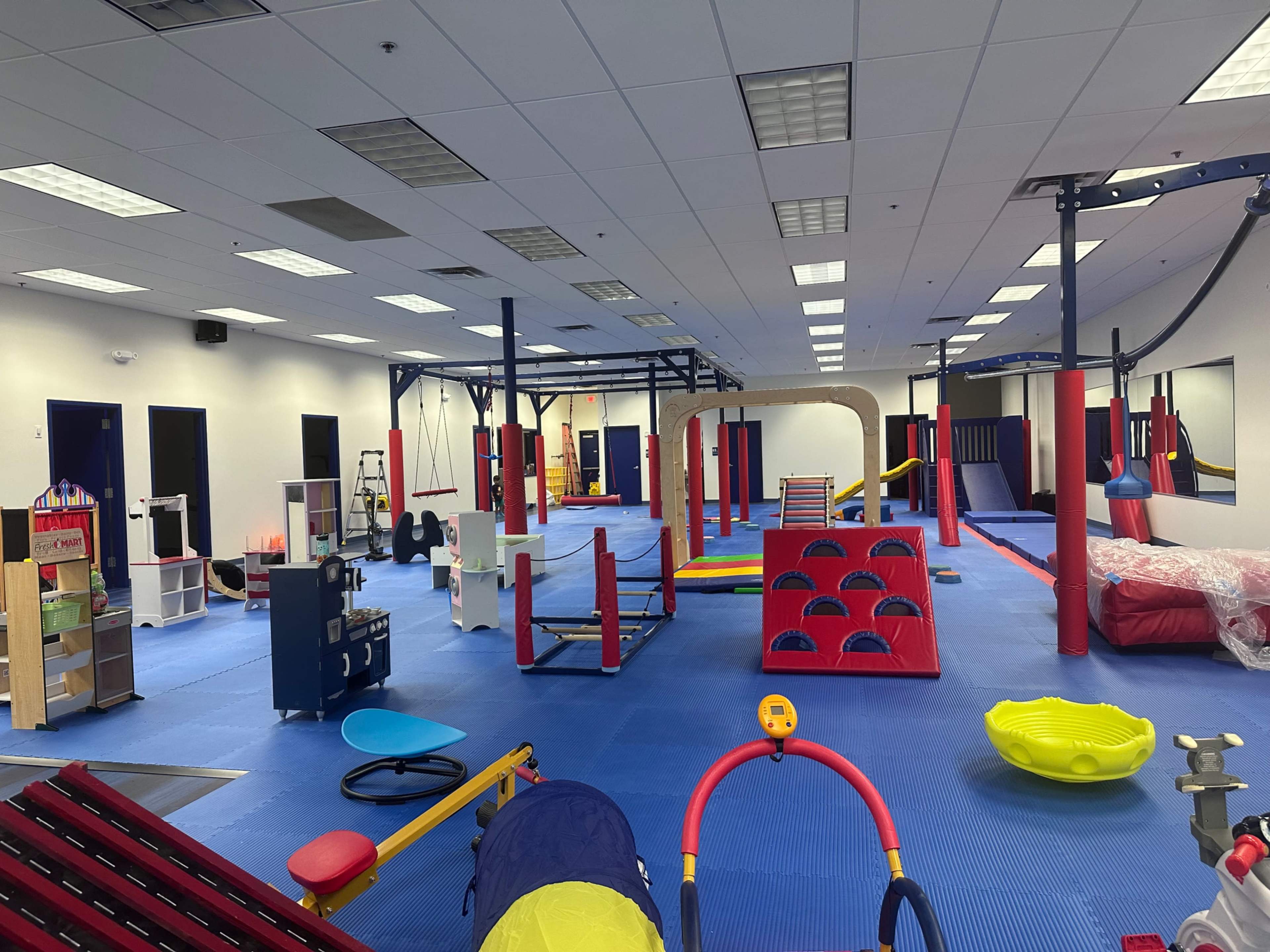 The image shows a brightly colored indoor play area equipped with various play structures, mats, and equipment arranged across a spacious room.