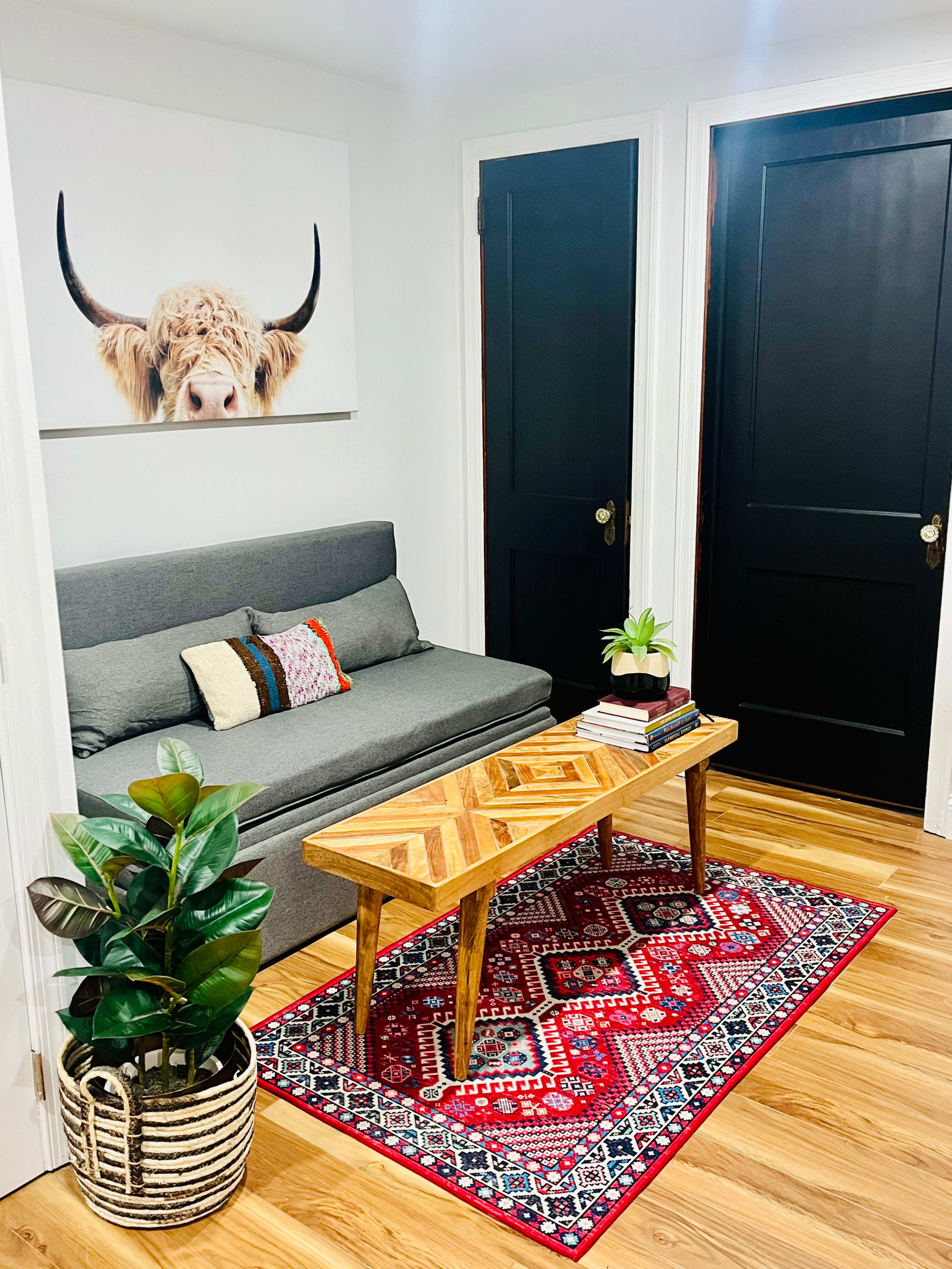 The image shows a cozy living room area featuring a gray sofa, a patterned rug, a wooden coffee table, and a plant beside two closed black doors and a wall art of a cow's face.