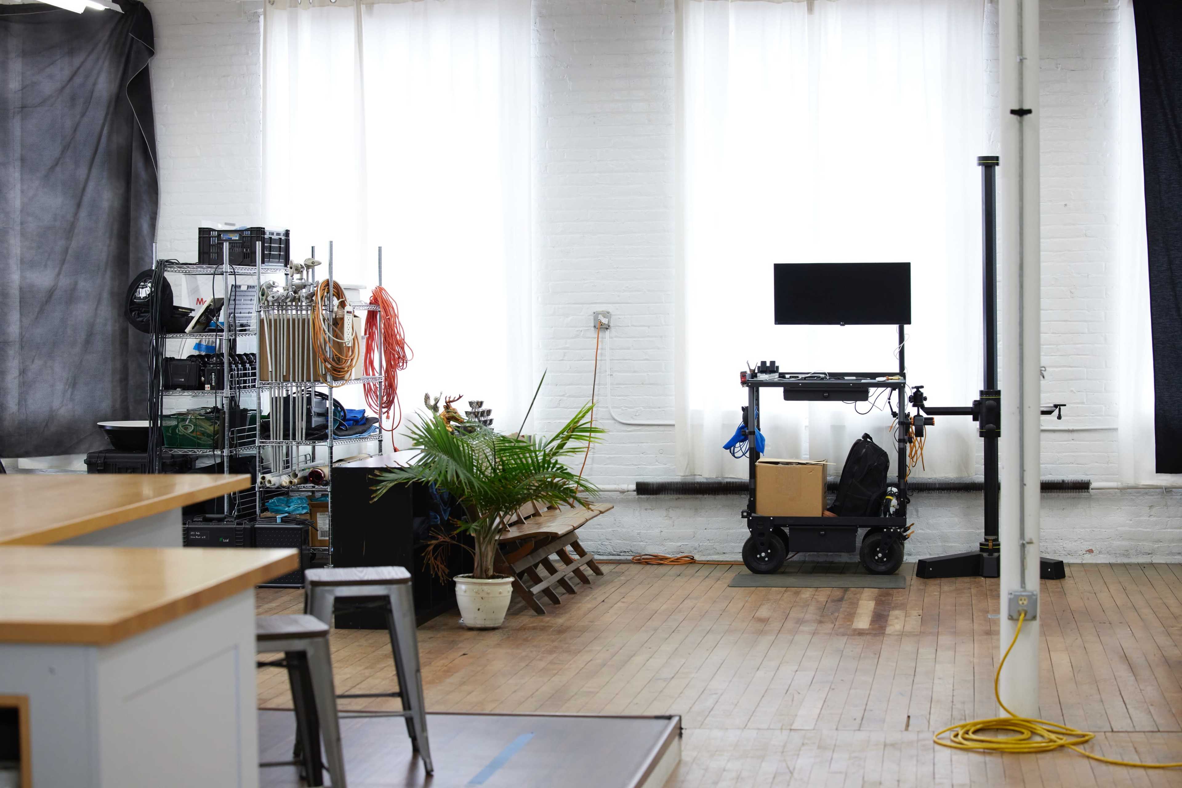 A spacious studio features large windows, a rack of equipment, a plant in a pot, and a cart with a television and various items.