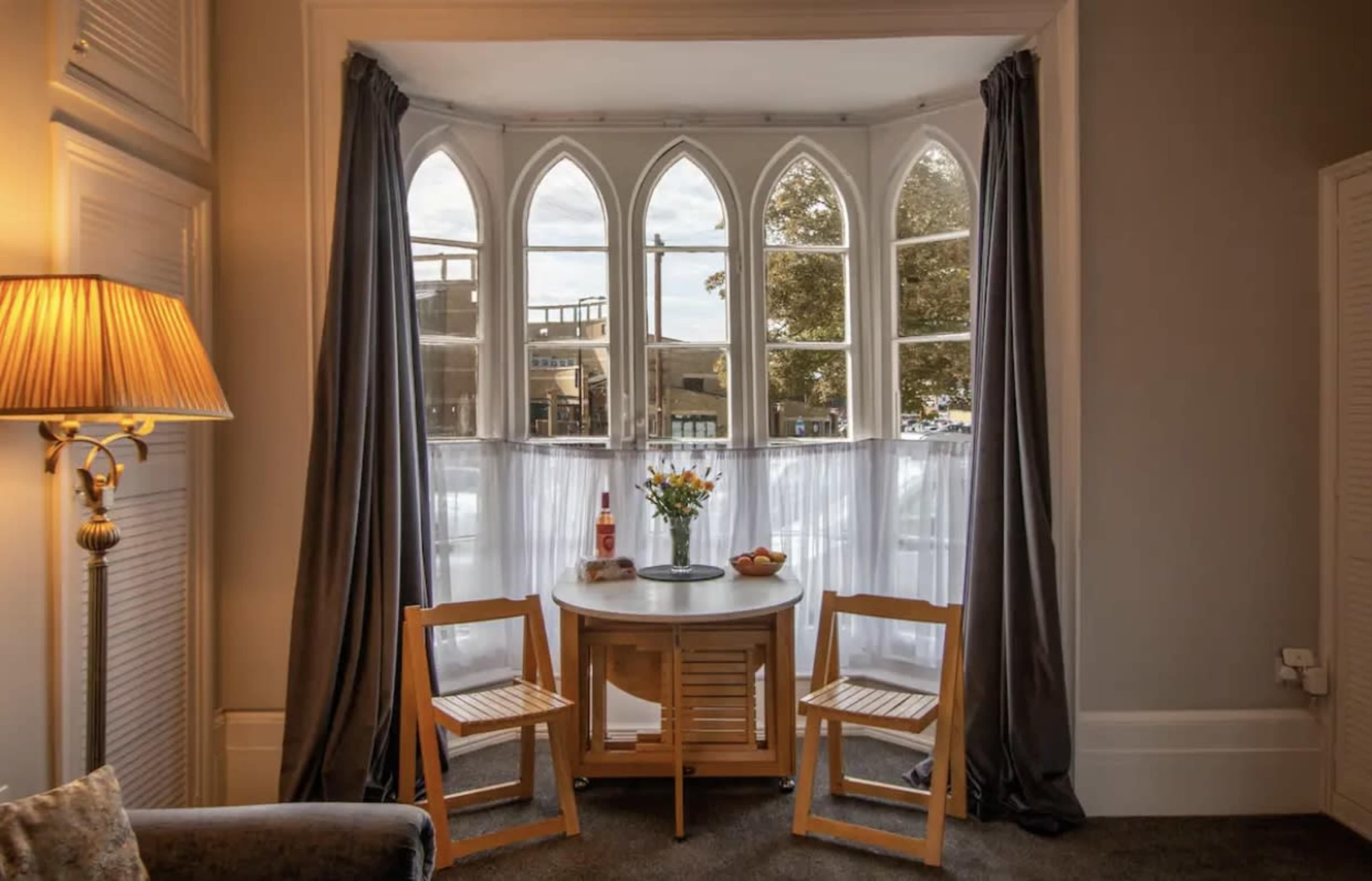 A small round table with two wooden chairs is set against a bay window framed by gray curtains, with a vase of flowers and a bowl of fruit on the table.