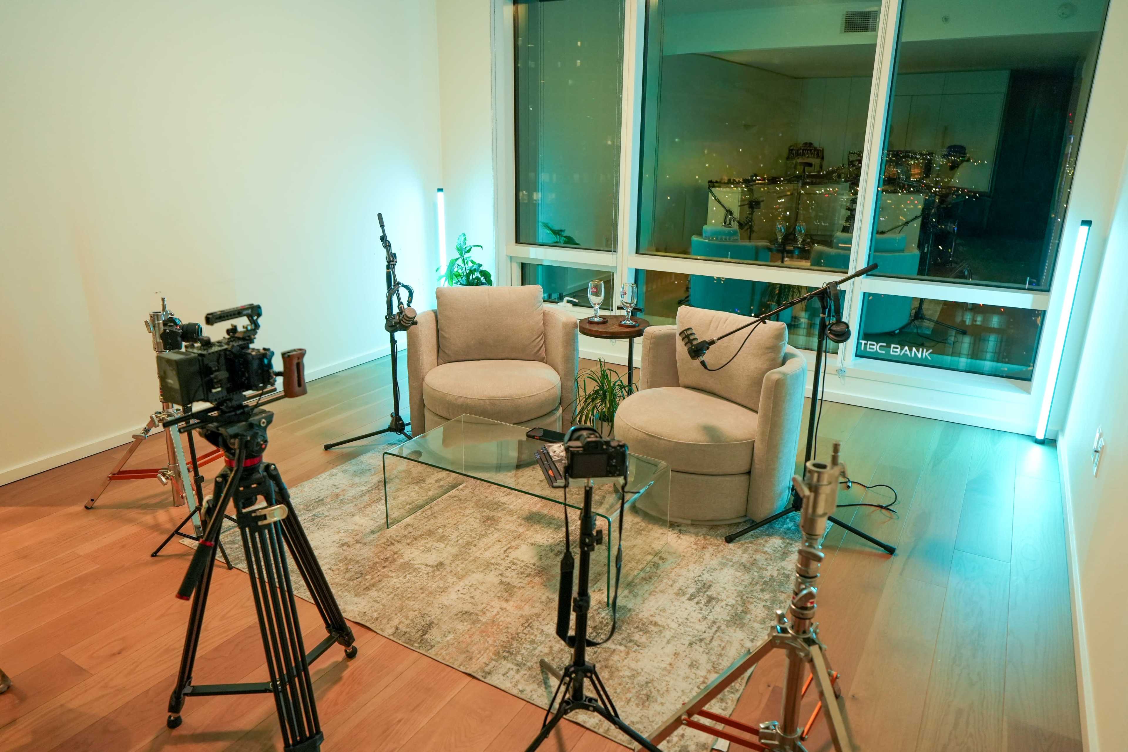 The image shows a modern sitting area with two light gray armchairs, a glass coffee table, and various camera equipment set up around it.