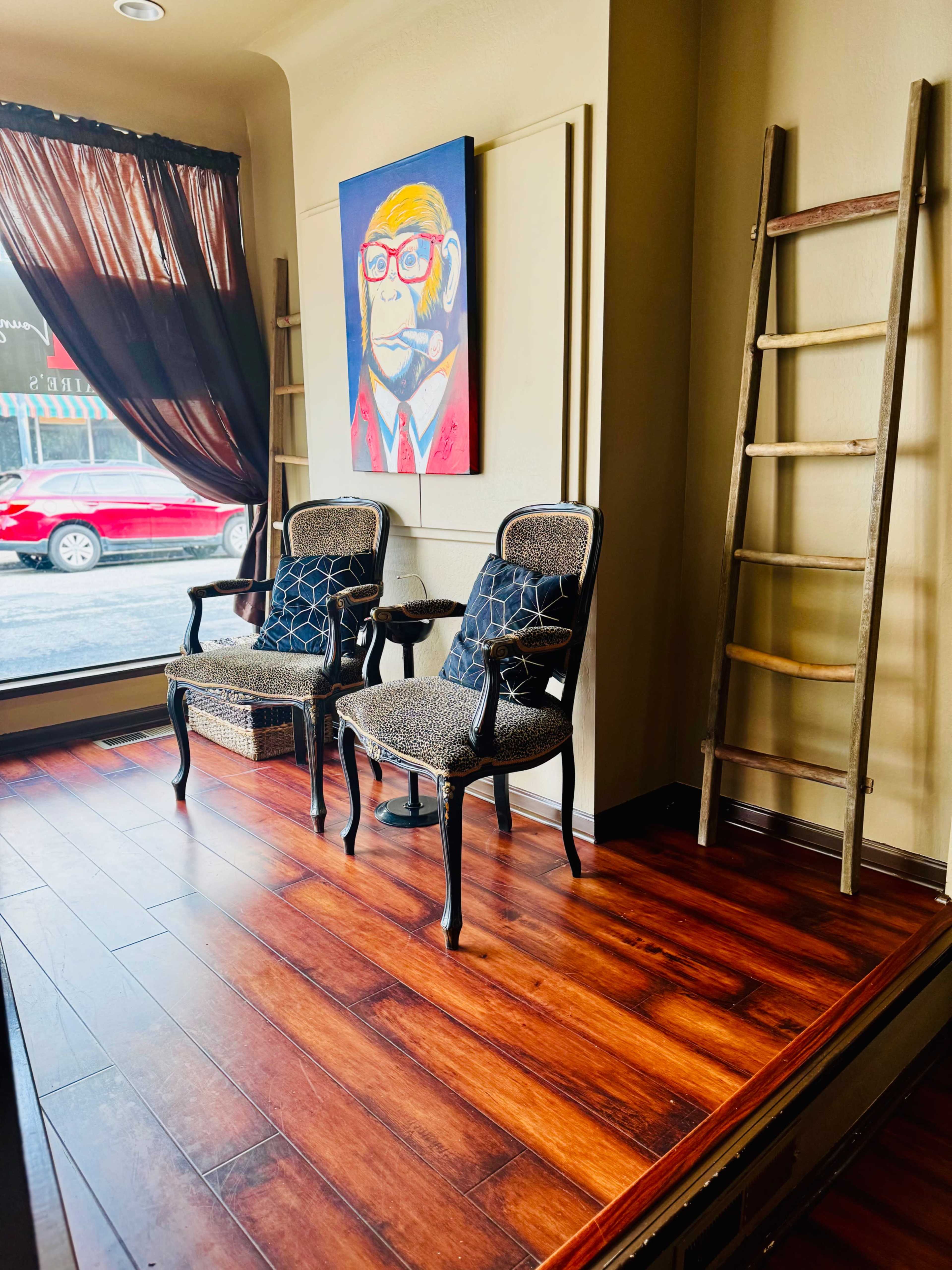 The image shows a small seating area with two patterned chairs beside a ladder and a colorful portrait hanging on the wall, illuminated by natural light coming through a window.