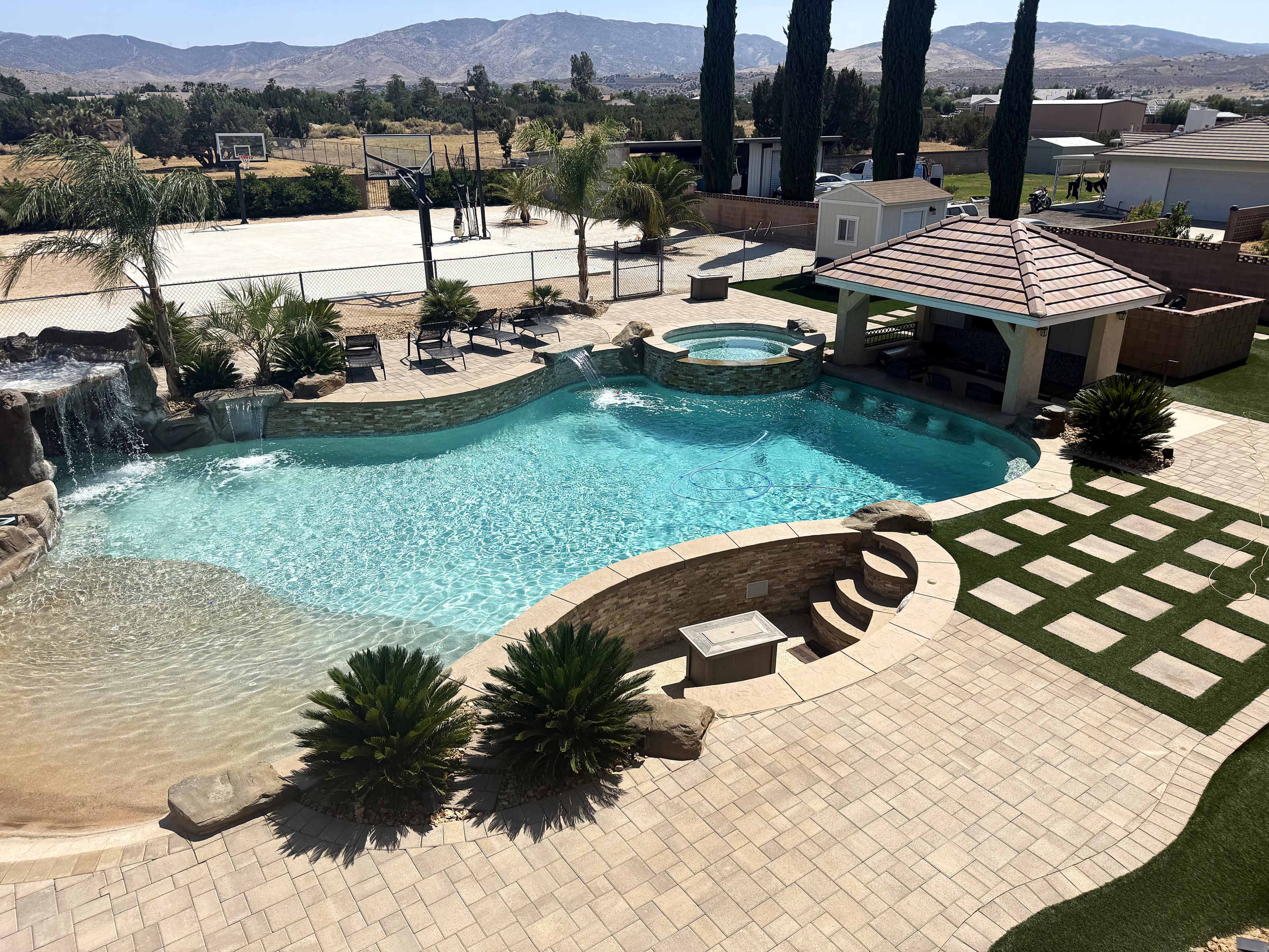 The image depicts a landscaped outdoor area featuring a swimming pool with a waterfall, spa, and patio, surrounded by palm trees and stone pathways.