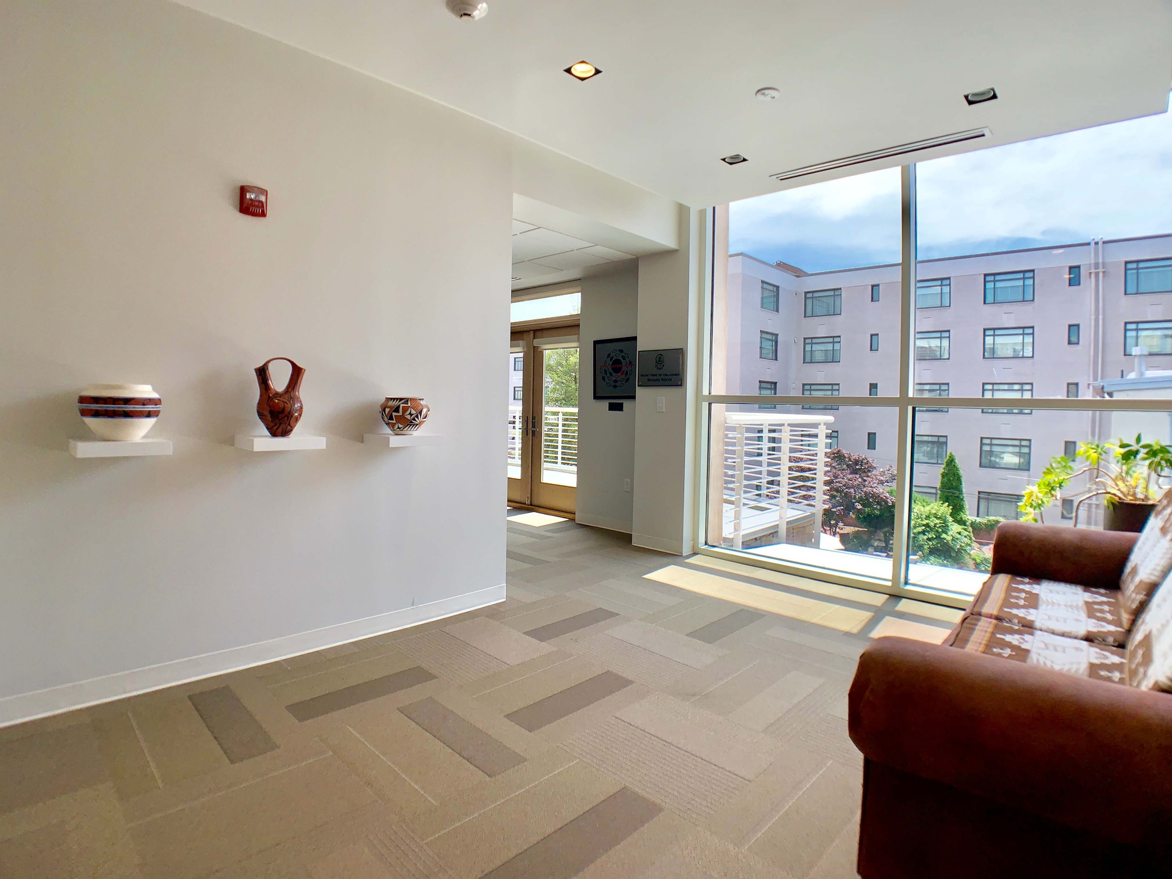 A well-lit interior space featuring a brown couch, a large window with a view of a garden, and three decorative pottery pieces displayed on white shelves.
