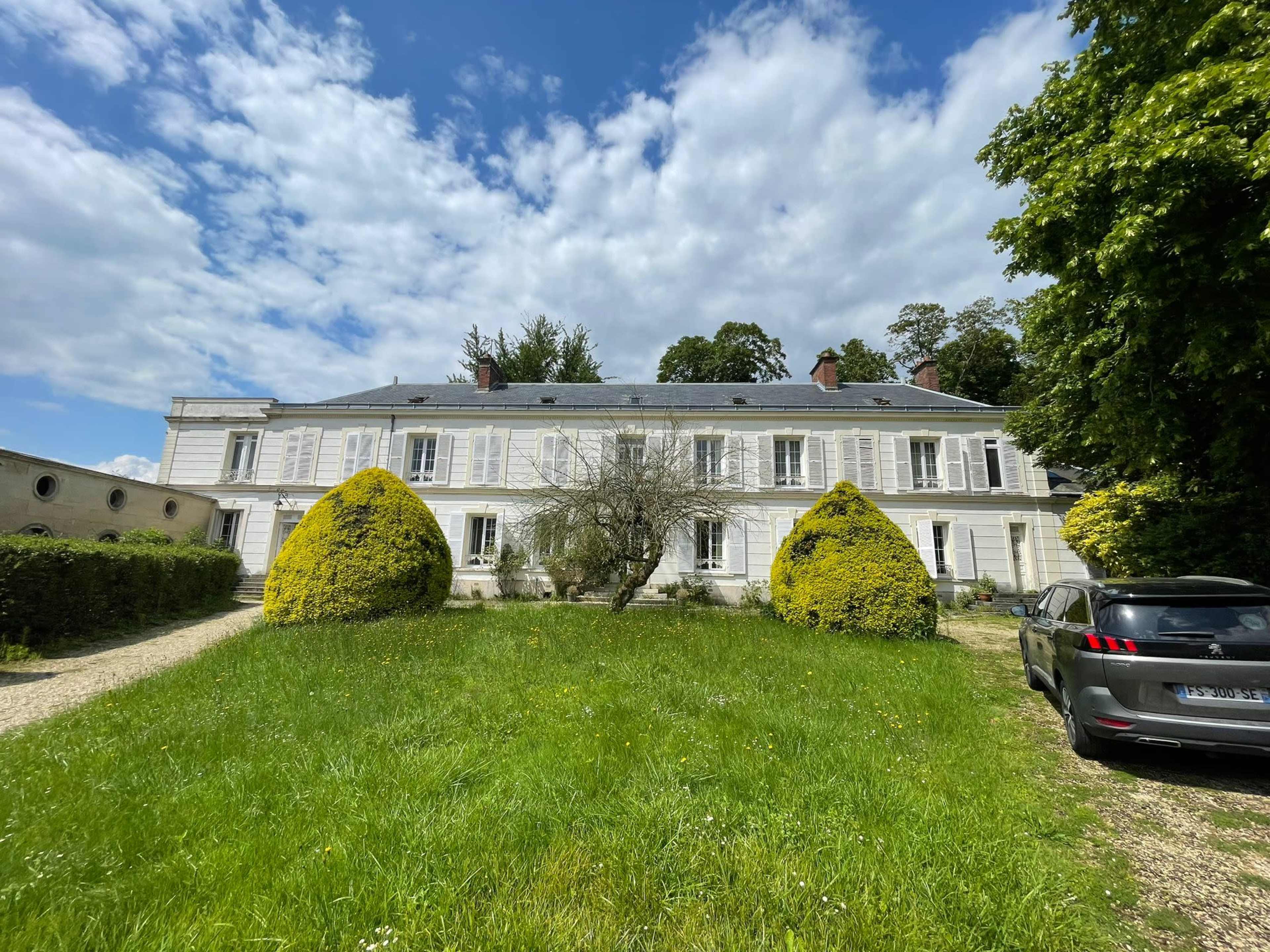 A large white house with multiple windows is set in a green landscape, featuring neatly trimmed shrubs and a gravel driveway.