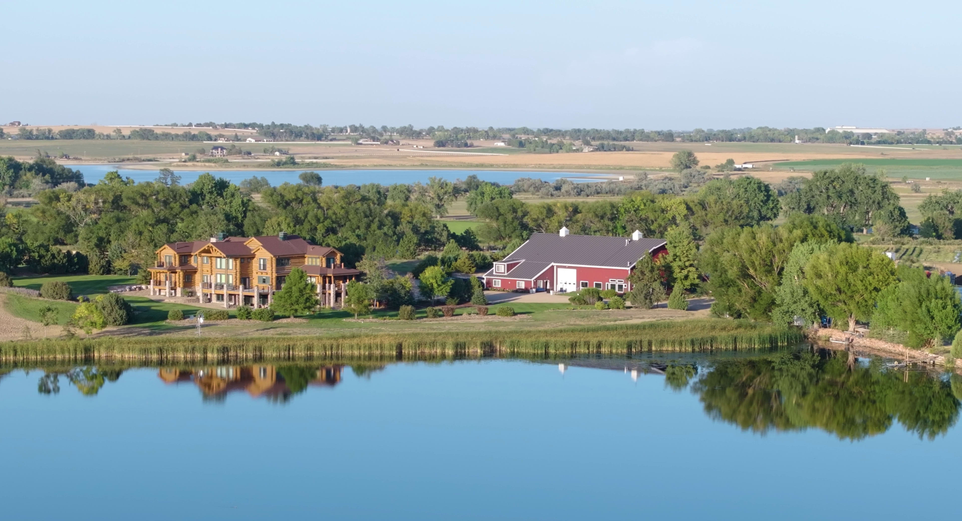 The image shows a serene landscape with a lake reflecting nearby houses and trees, set against rolling green fields under a clear sky.